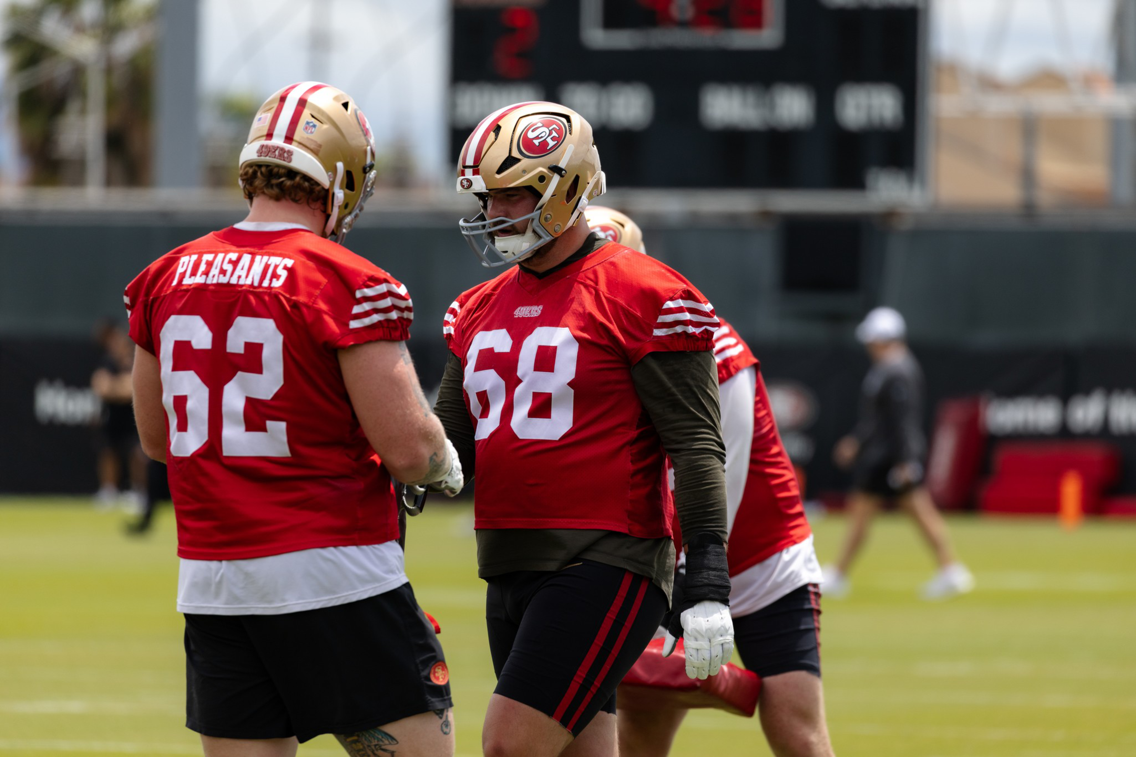 Two football players wearing red San Francisco 49ers jerseys, numbered 62 and 68, stand on a grassy field during practice.