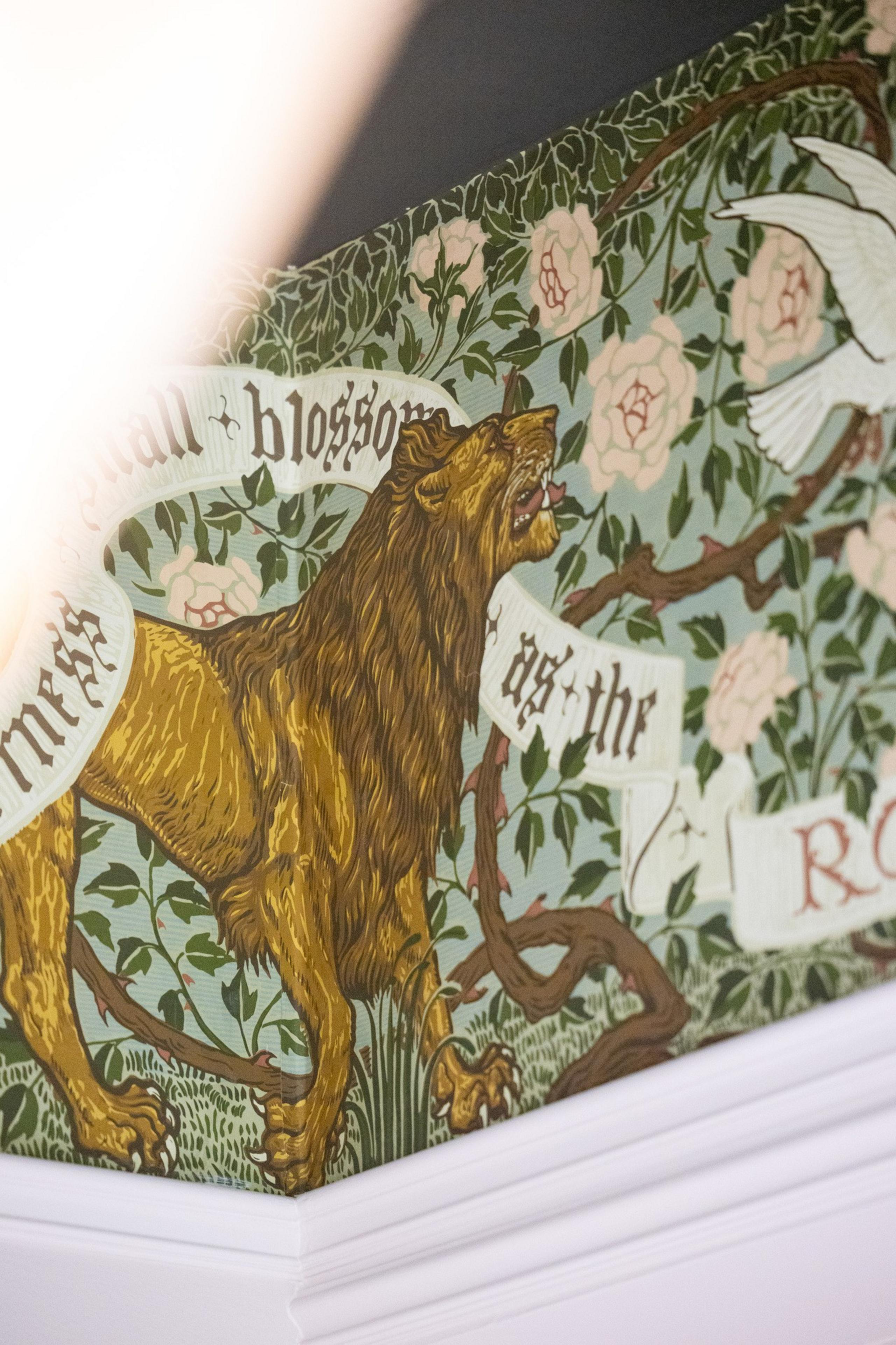 The image shows a detailed mural of a roaring lion surrounded by foliage and pink flowers. White birds fly nearby, and there is text intertwined with the scene.