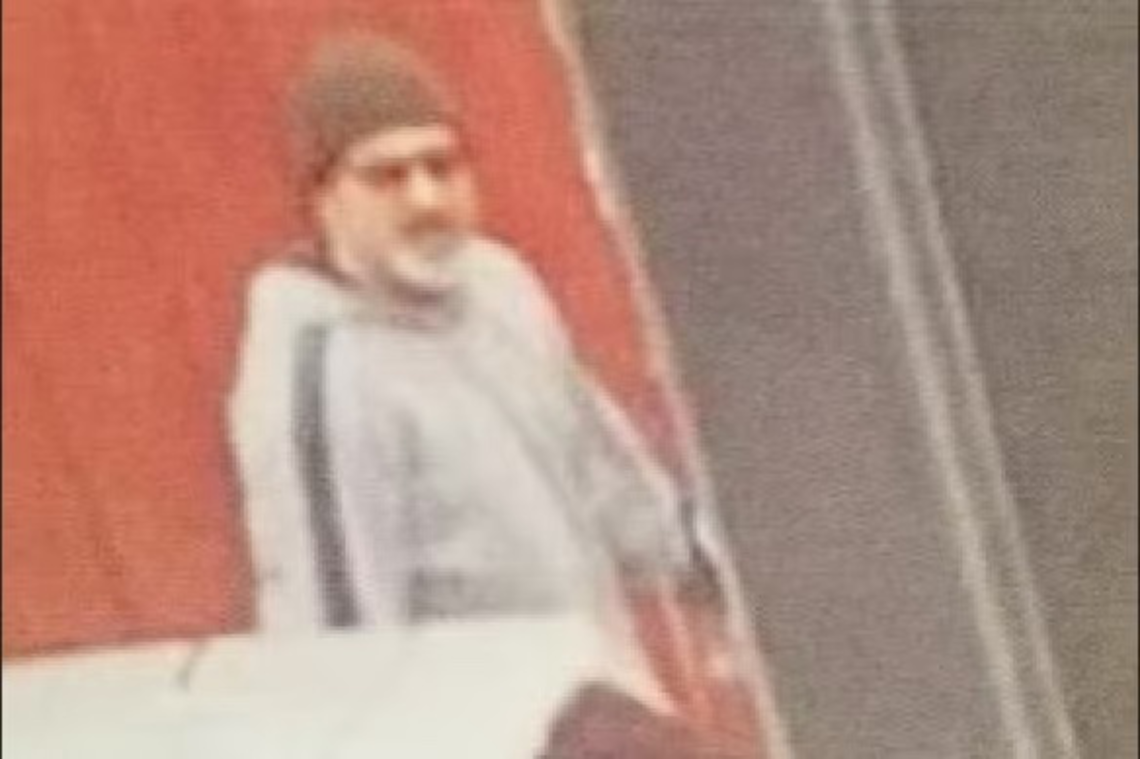 A blurry image of a person seated, wearing a cap and a light-colored outfit, with a red backdrop.