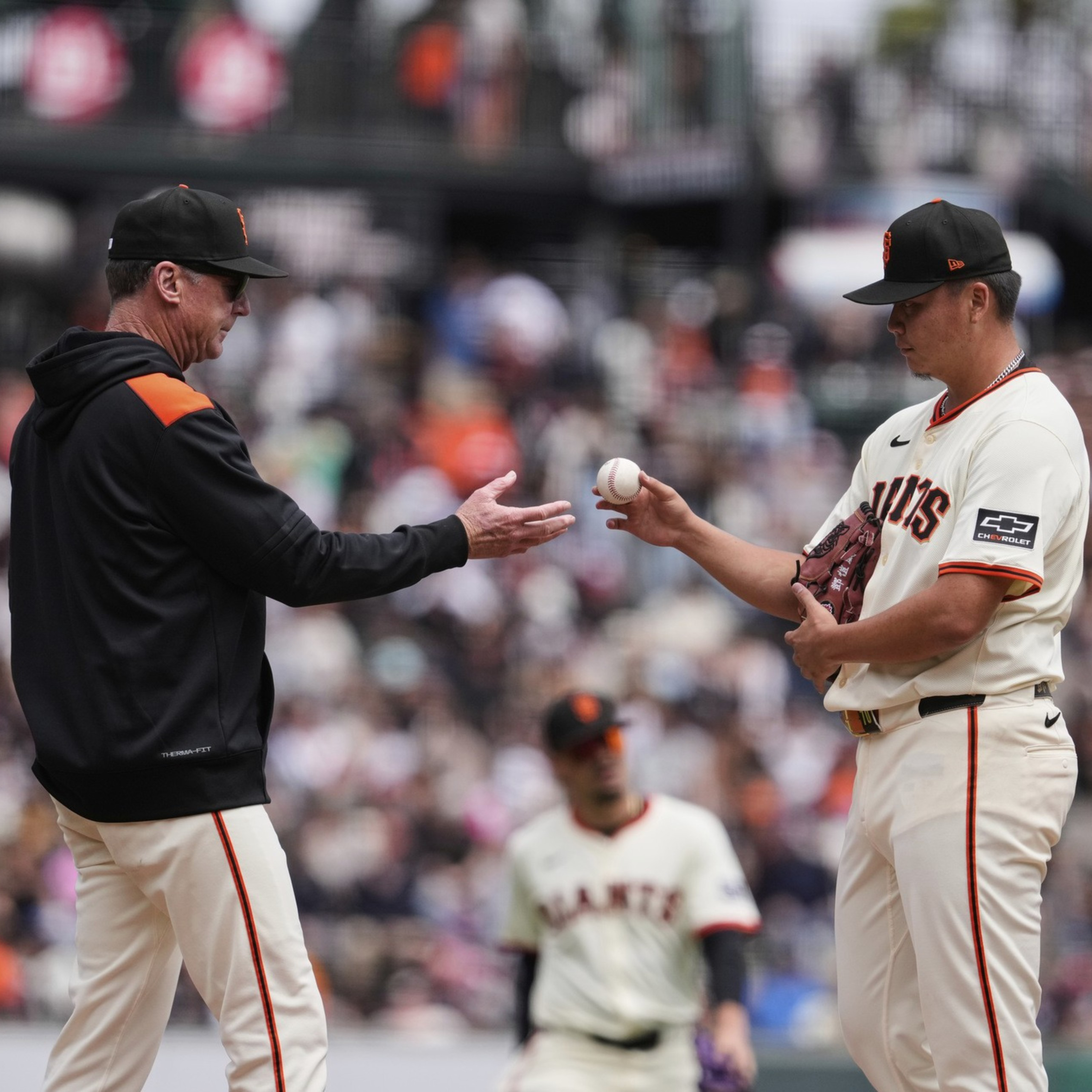 A baseball coach wearing a black jacket and cap hands a ball to a player in a white Giants uniform while two other players stand nearby.