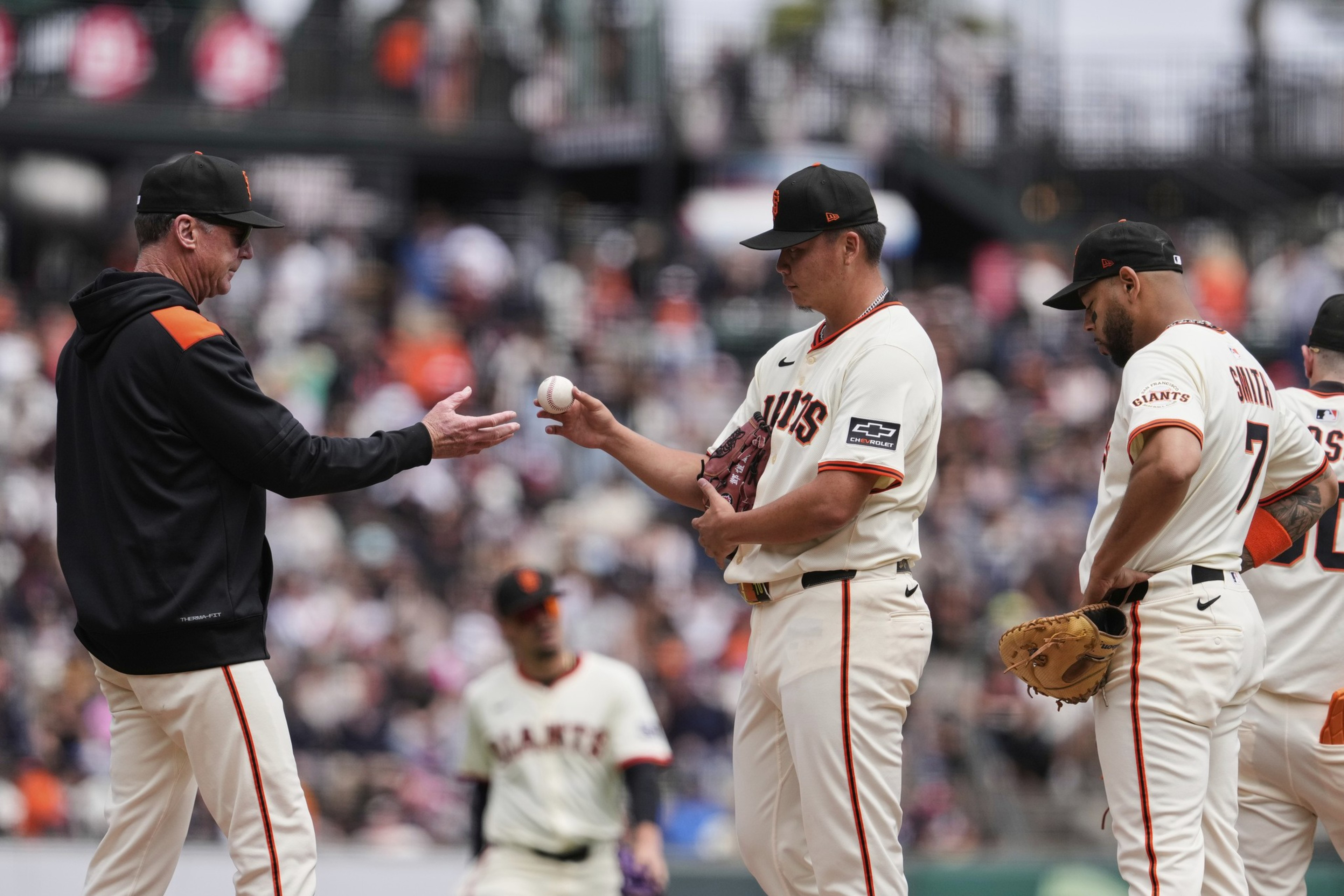 A baseball coach wearing a black jacket and cap hands a ball to a player in a white Giants uniform while two other players stand nearby.
