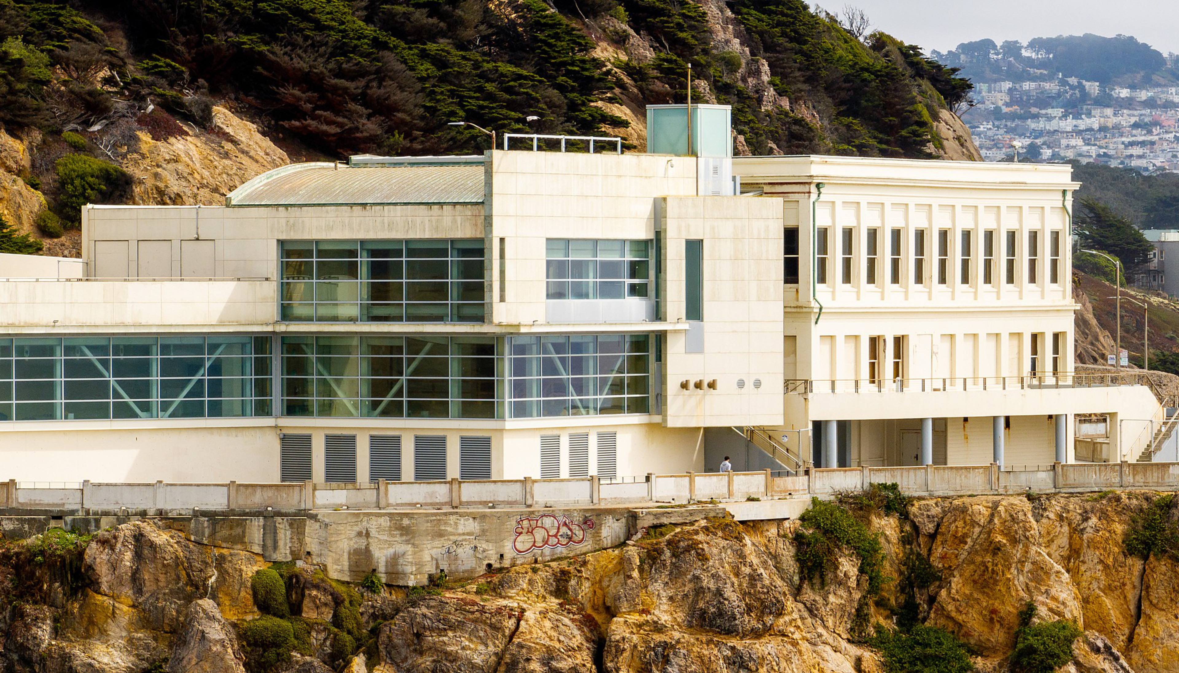 A large white building with many windows sits atop rocky cliffs with greenery behind and a distant cityscape visible.