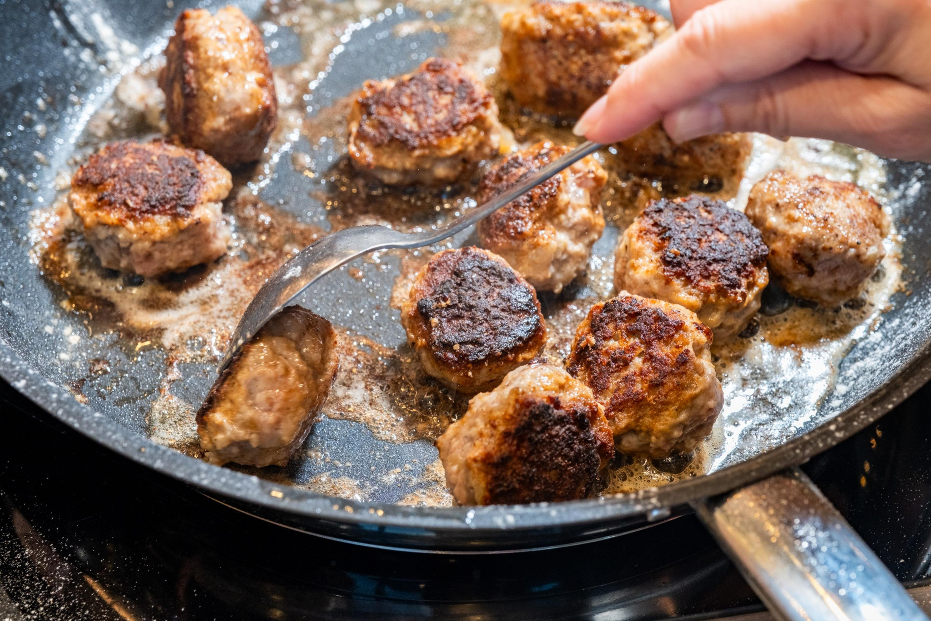 A hand uses a spoon to turn browned meatballs sizzling in a frying pan, surrounded by melted oil or butter.