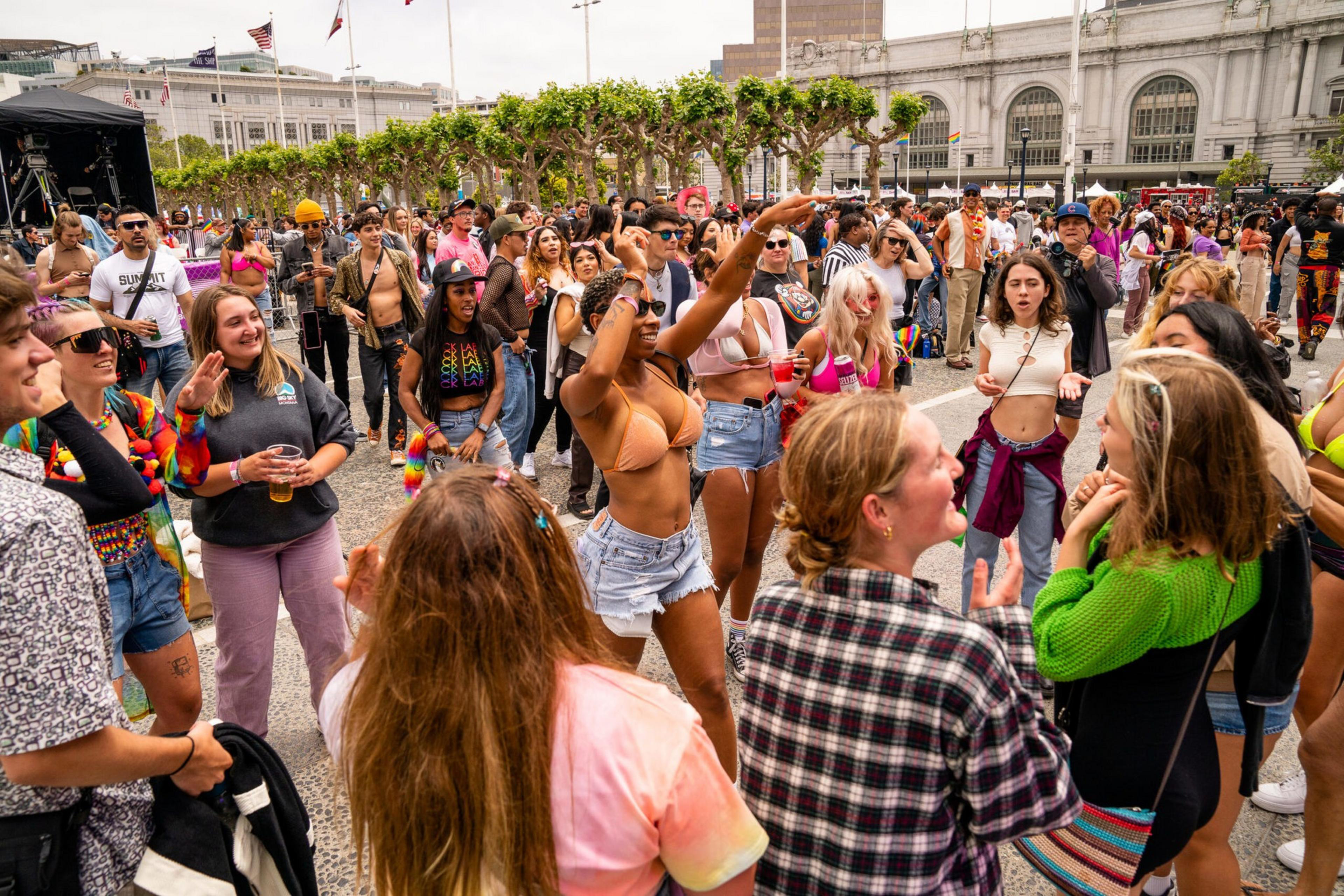 A diverse crowd dances and socializes outdoors at a lively event with colorful outfits and a historic building in the background.