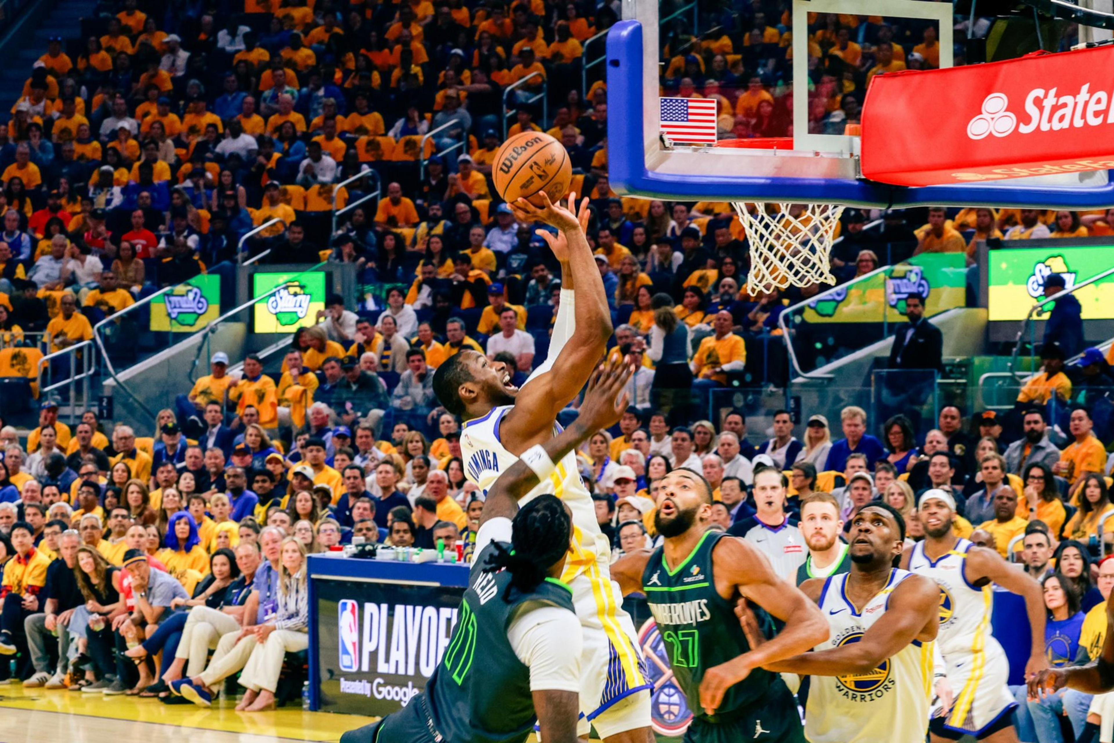 A basketball player in a white and blue uniform leaps toward the hoop to score, surrounded by opposing players in green. The crowd watches intently.