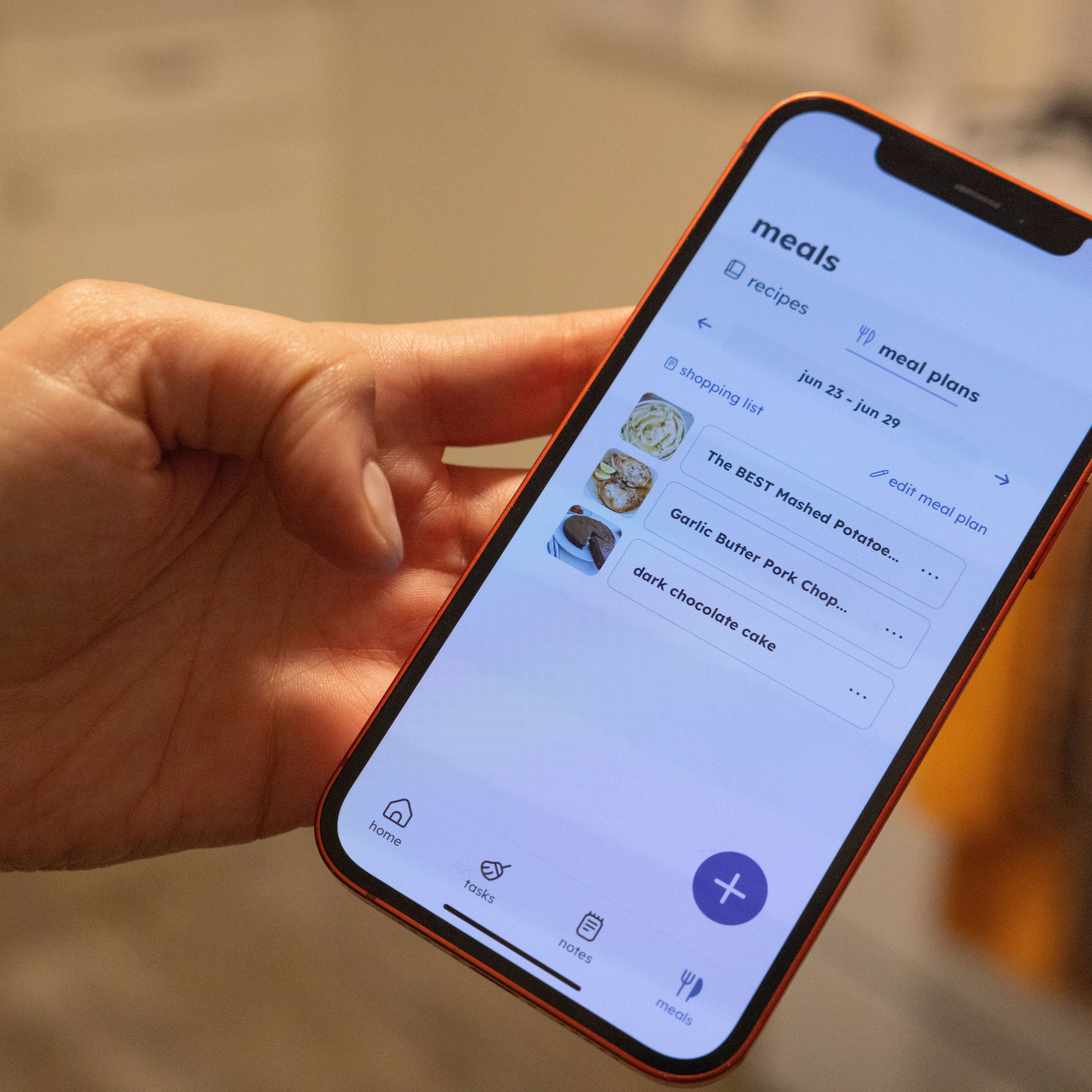 A hand holds a smartphone displaying a meal planning app. The screen shows recipes and meal plans, listing dishes like mashed potatoes, pork chops, and chocolate cake.