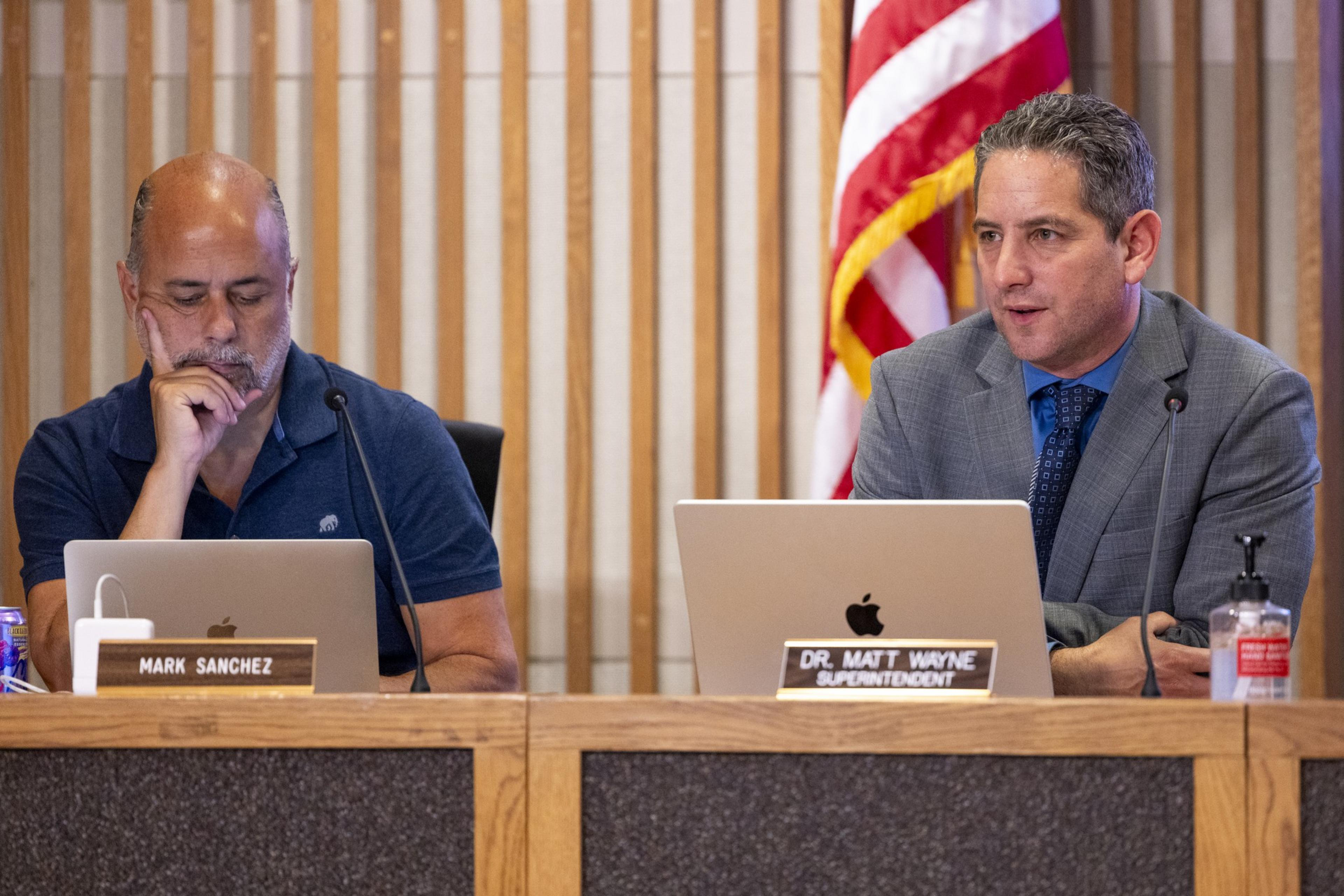 Two men sit at a table with laptops and nameplates. One is focused, and the other is speaking. There's a U.S. flag in the background.
