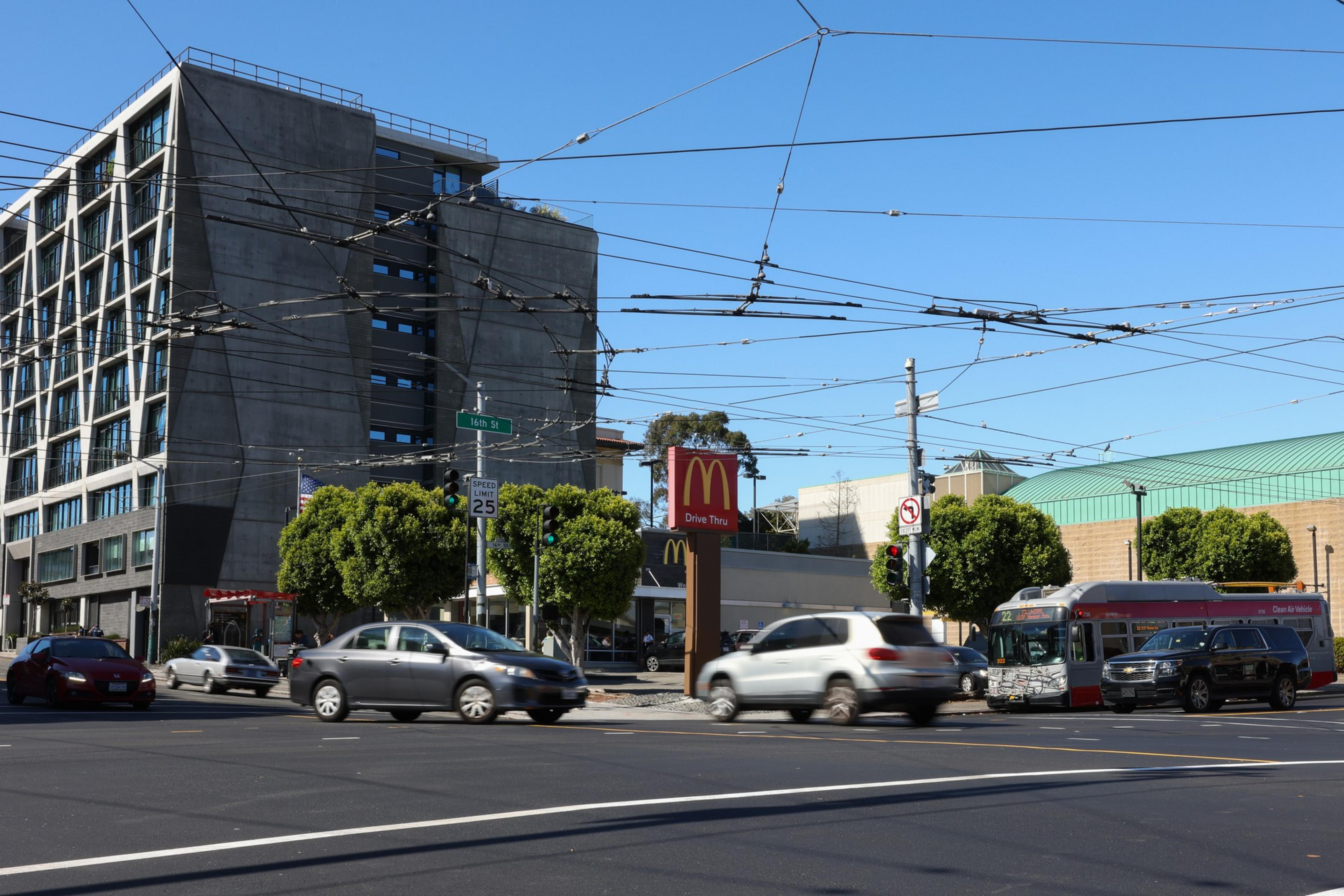 A busy urban intersection includes cars, a bus, a McDonald's drive-thru sign, a modern multi-story building, and overhead trolleybus wires.