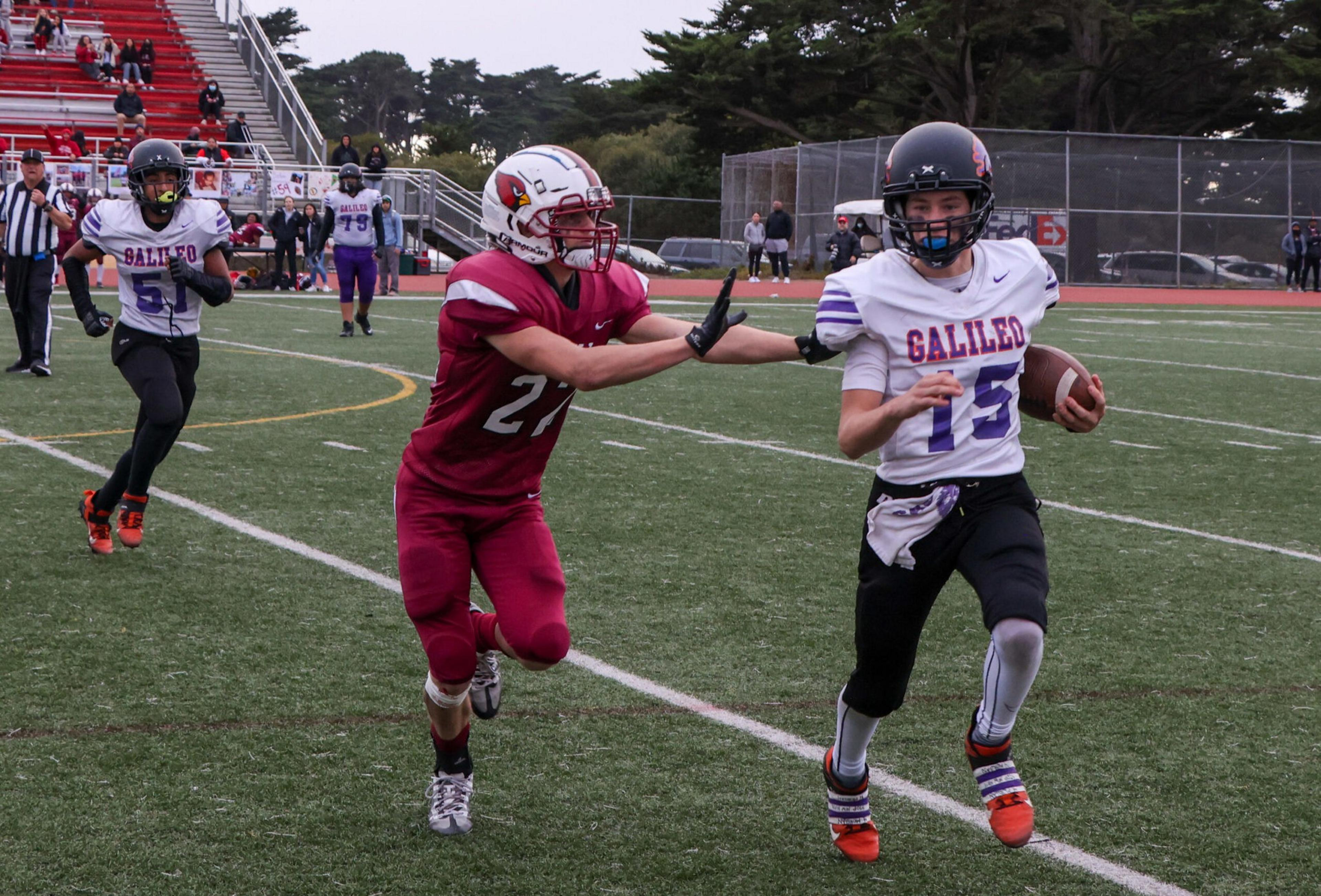 A football player in a white and purple jersey runs with the ball while a player in a red uniform reaches out to block him on a green field.
