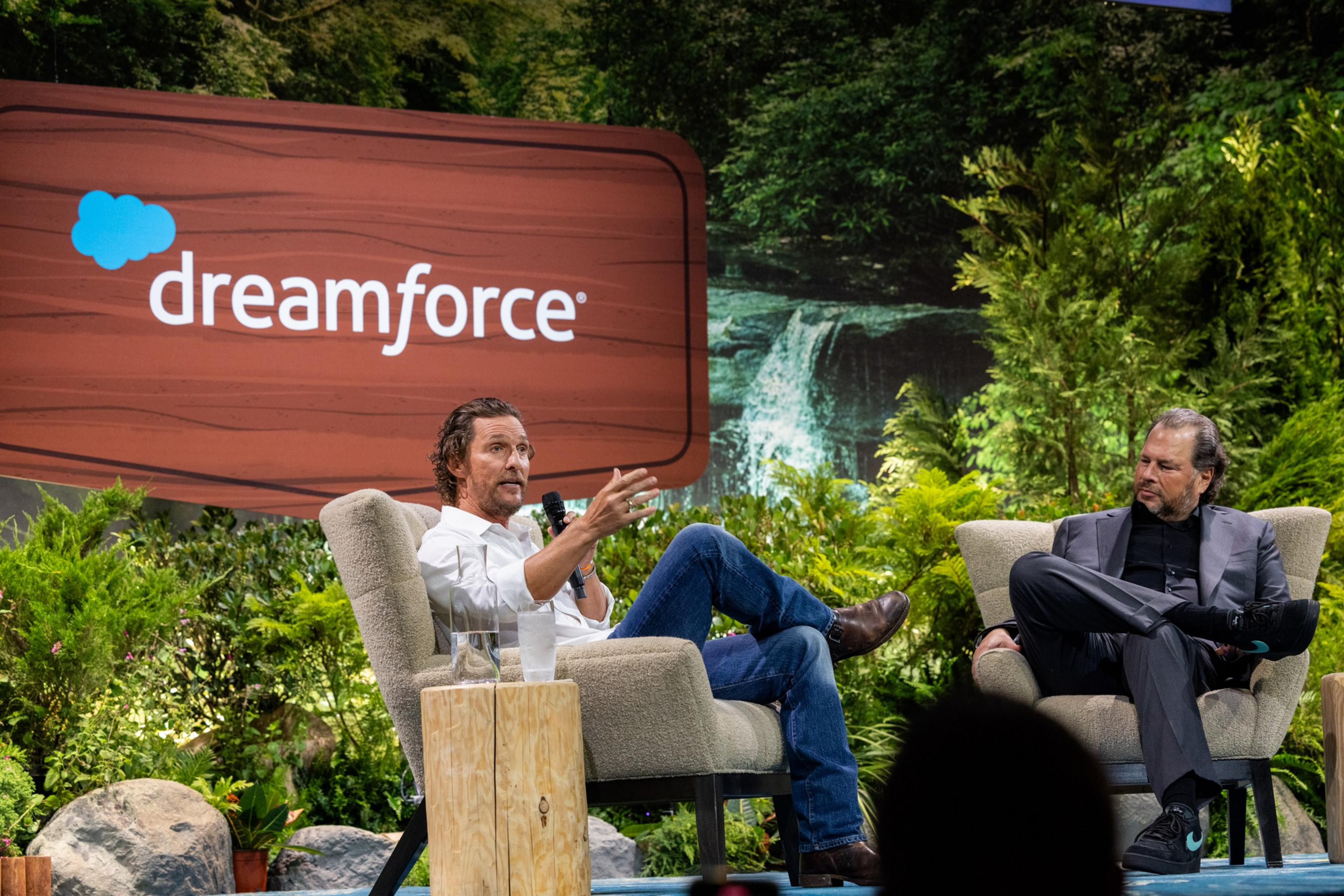 Two men sit in armchairs onstage at Dreamforce, one speaking into a microphone while the other listens, surrounded by lush greenery and a waterfall backdrop.