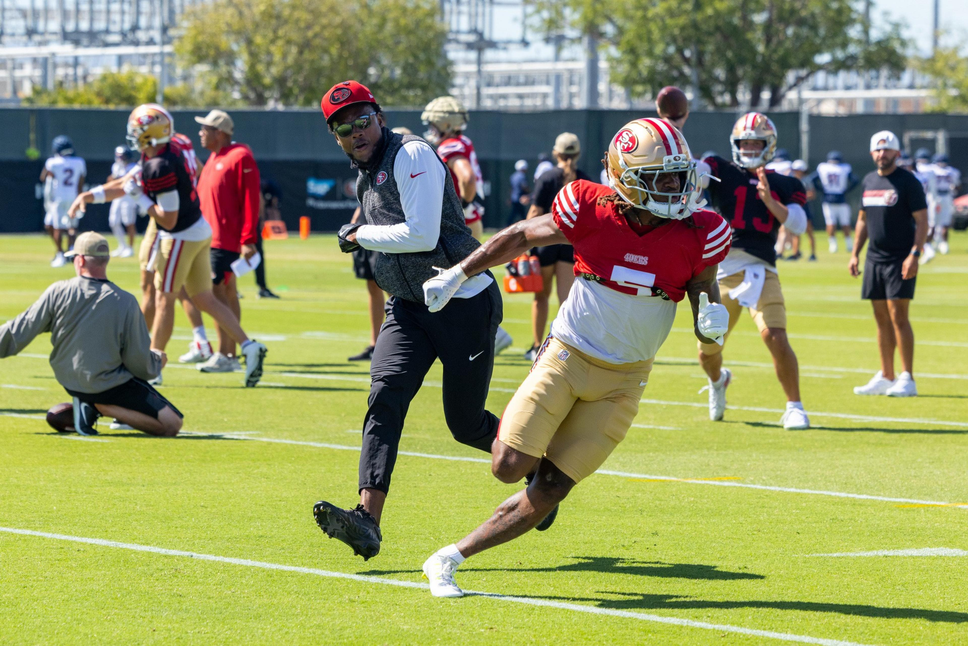 A football player in a red and gold San Francisco 49ers uniform runs on a grassy field while a coach in black pants and a red cap watches closely.