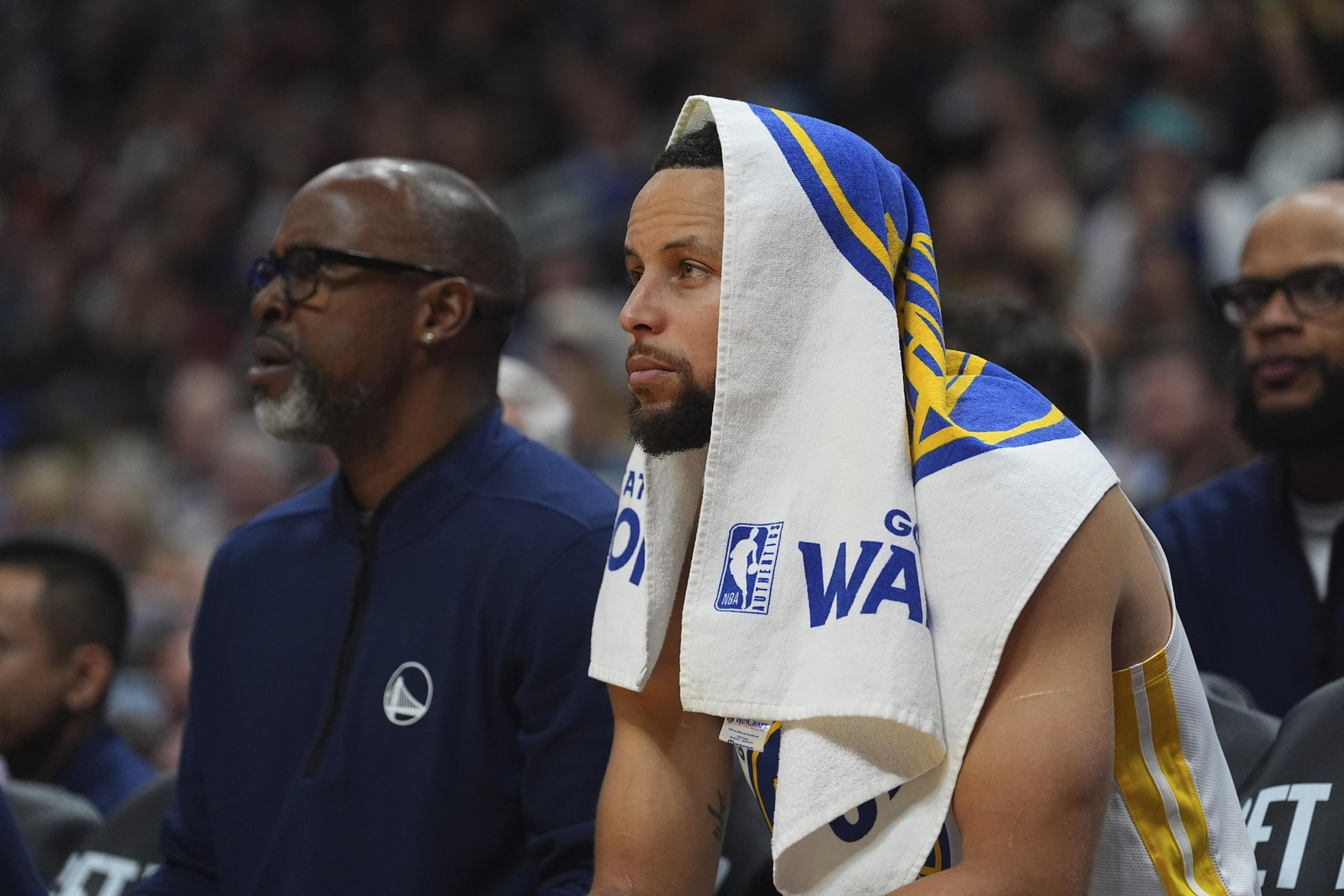 A basketball player sits on the bench with a towel draped over their head. Another person in sports attire sits beside them. The setting appears to be a game.
