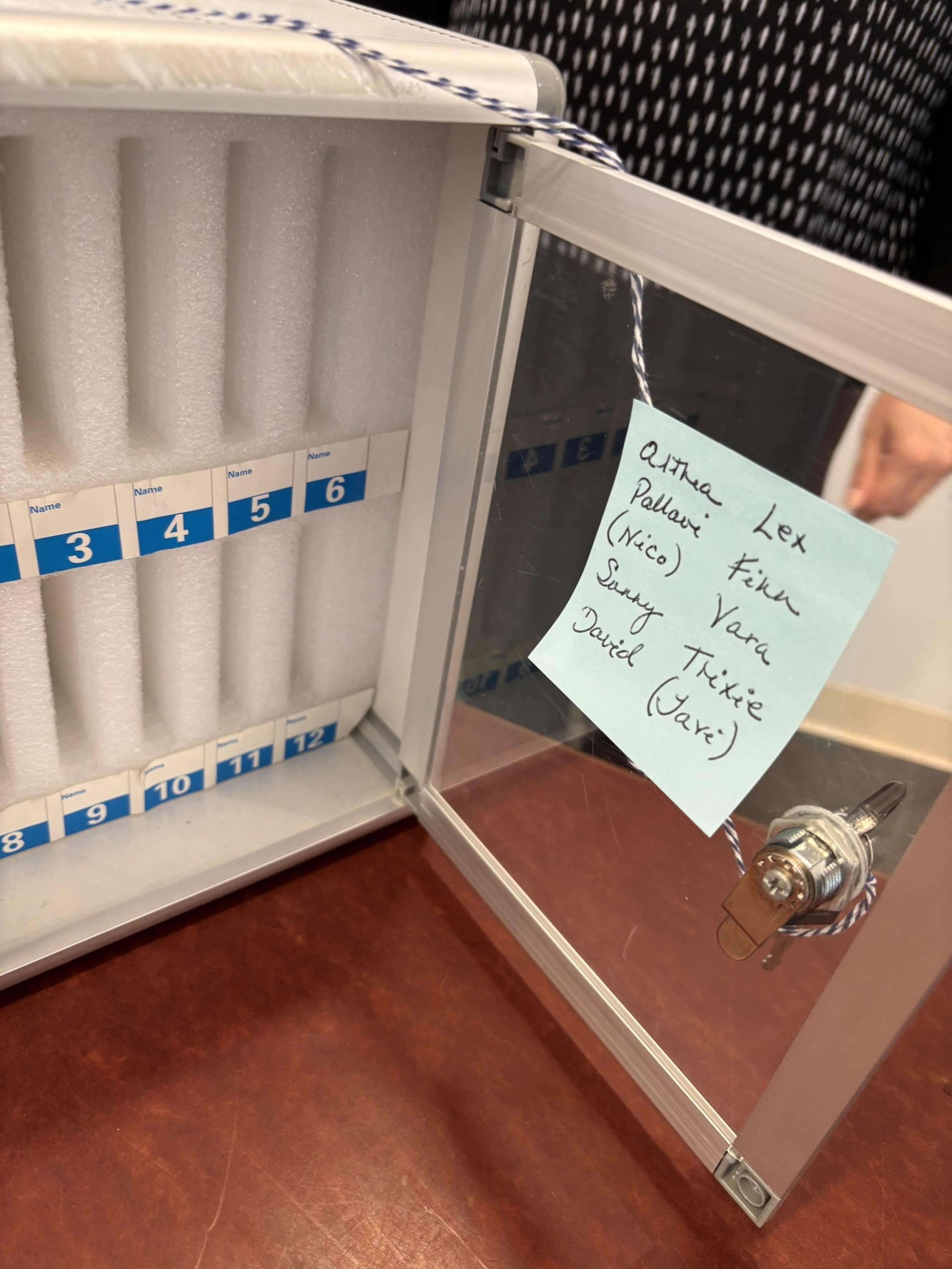 A small white storage box with numbered slots from 1 to 12, a locked transparent door, and a blue sticky note with handwritten names inside the door.