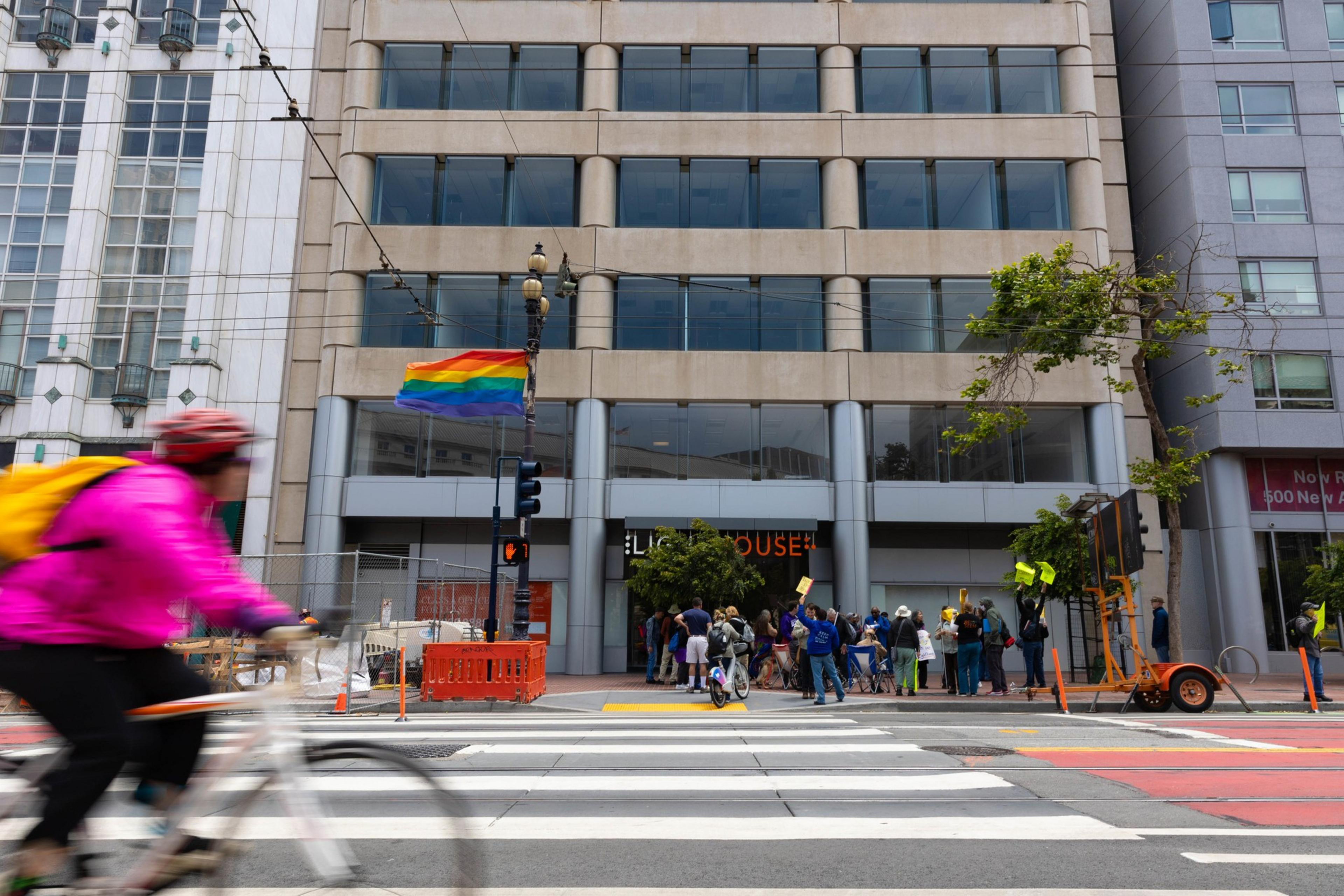 A cyclist in a bright pink jacket rides past a building with a rainbow flag. On the other side of the street, a group of people hold protest signs in front of LightHouse.