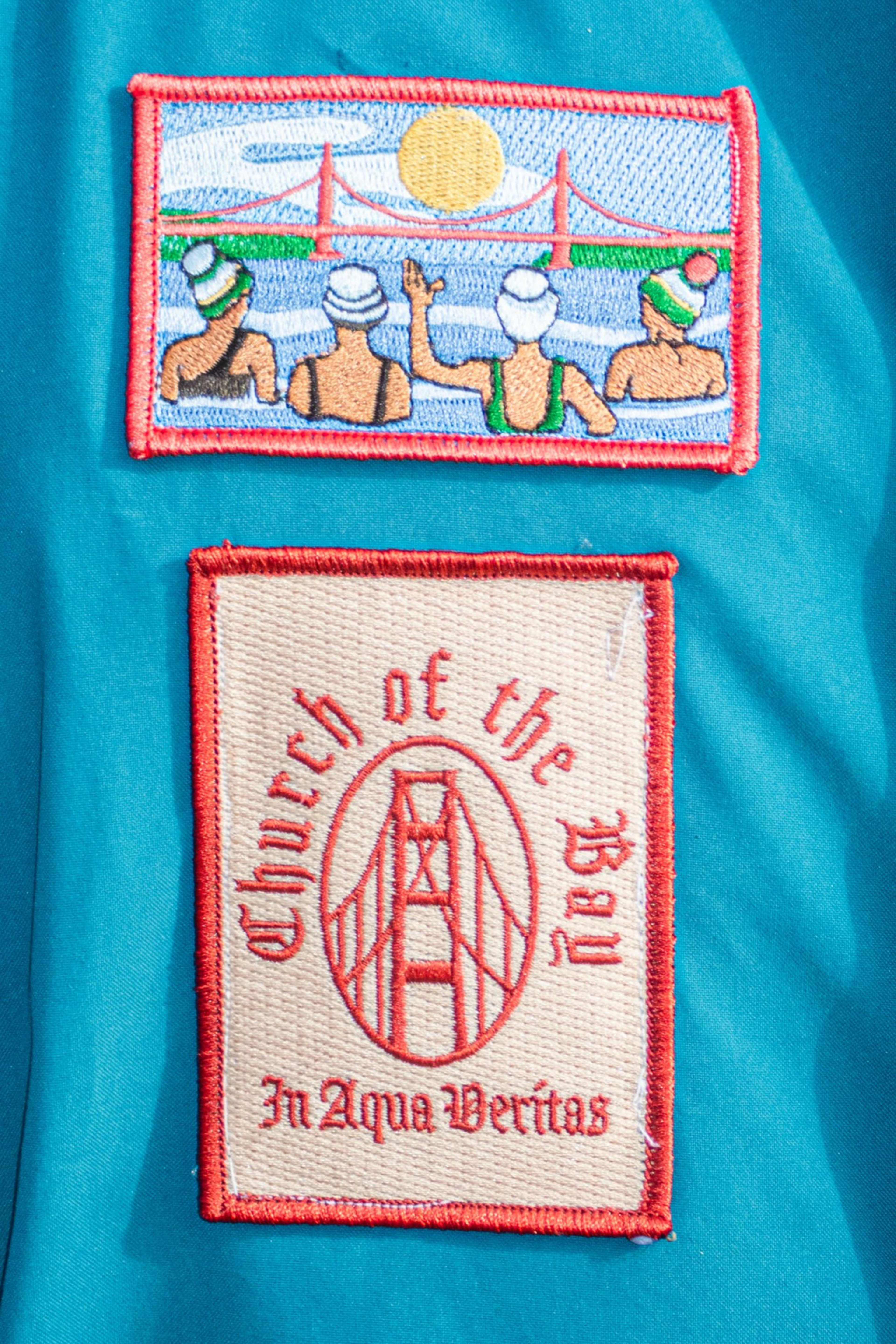 The image shows two embroidered patches. The top patch depicts swimmers and a bridge under the sun. The bottom patch reads &quot;Church of the Bay&quot; and &quot;In Aqua Veritas.&quot;