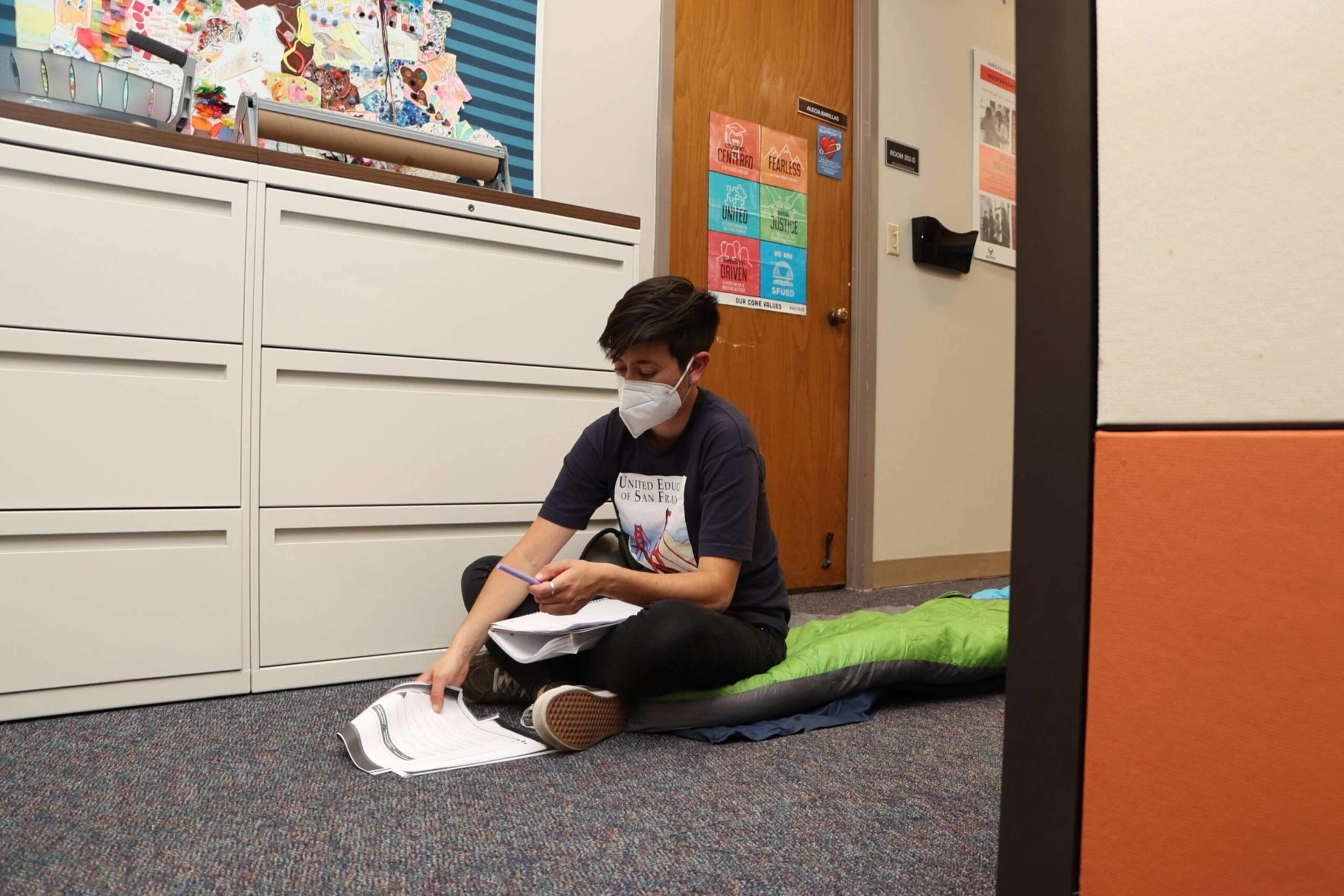 A person wearing a mask, sitting cross-legged on a sleeping bag on the floor, writing on papers inside a room with white cabinets and a wooden door.