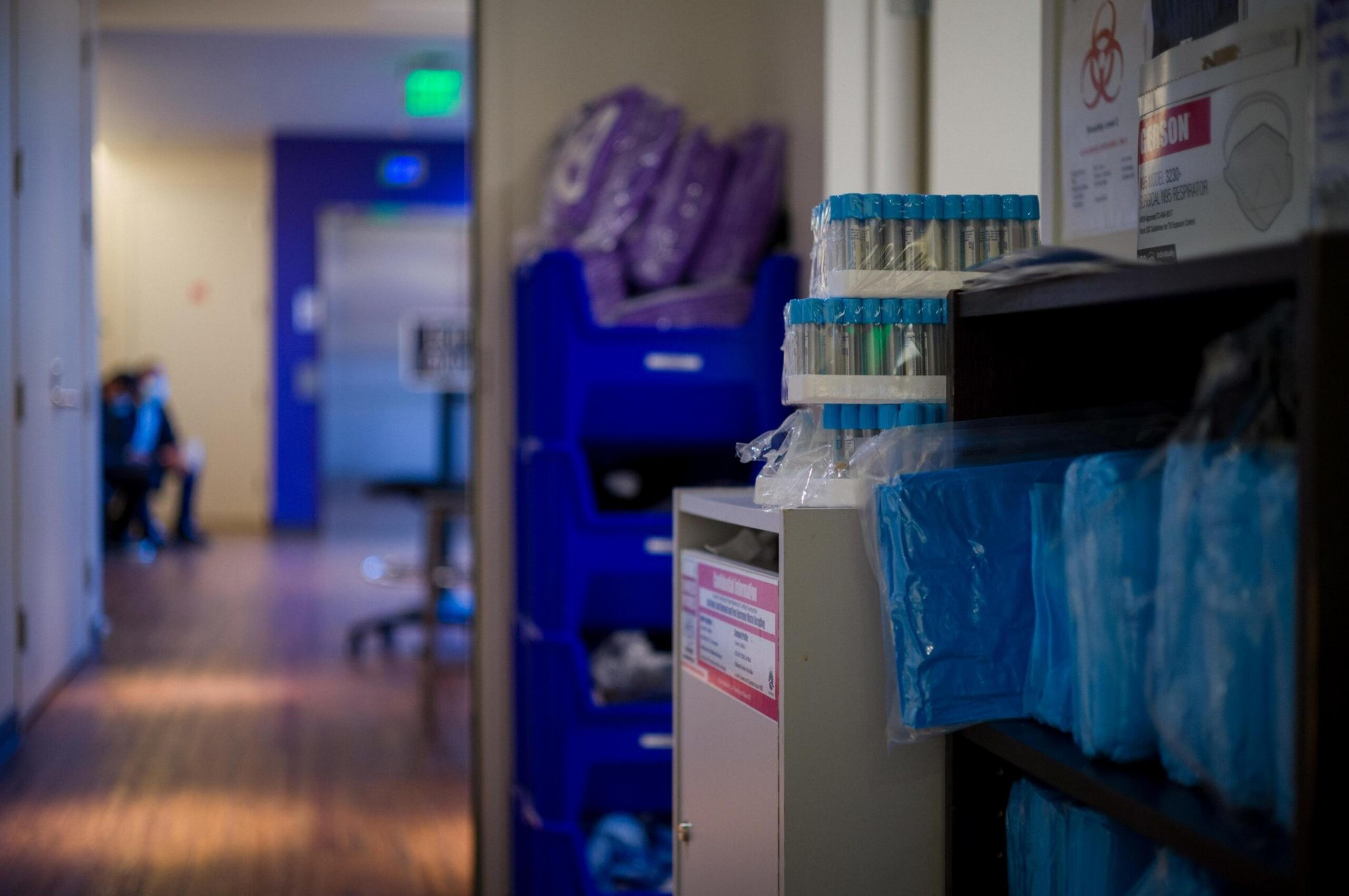 Shelves hold medical supplies like test tubes and gowns, with a hallway featuring a person sitting in blue scrubs at the far end.