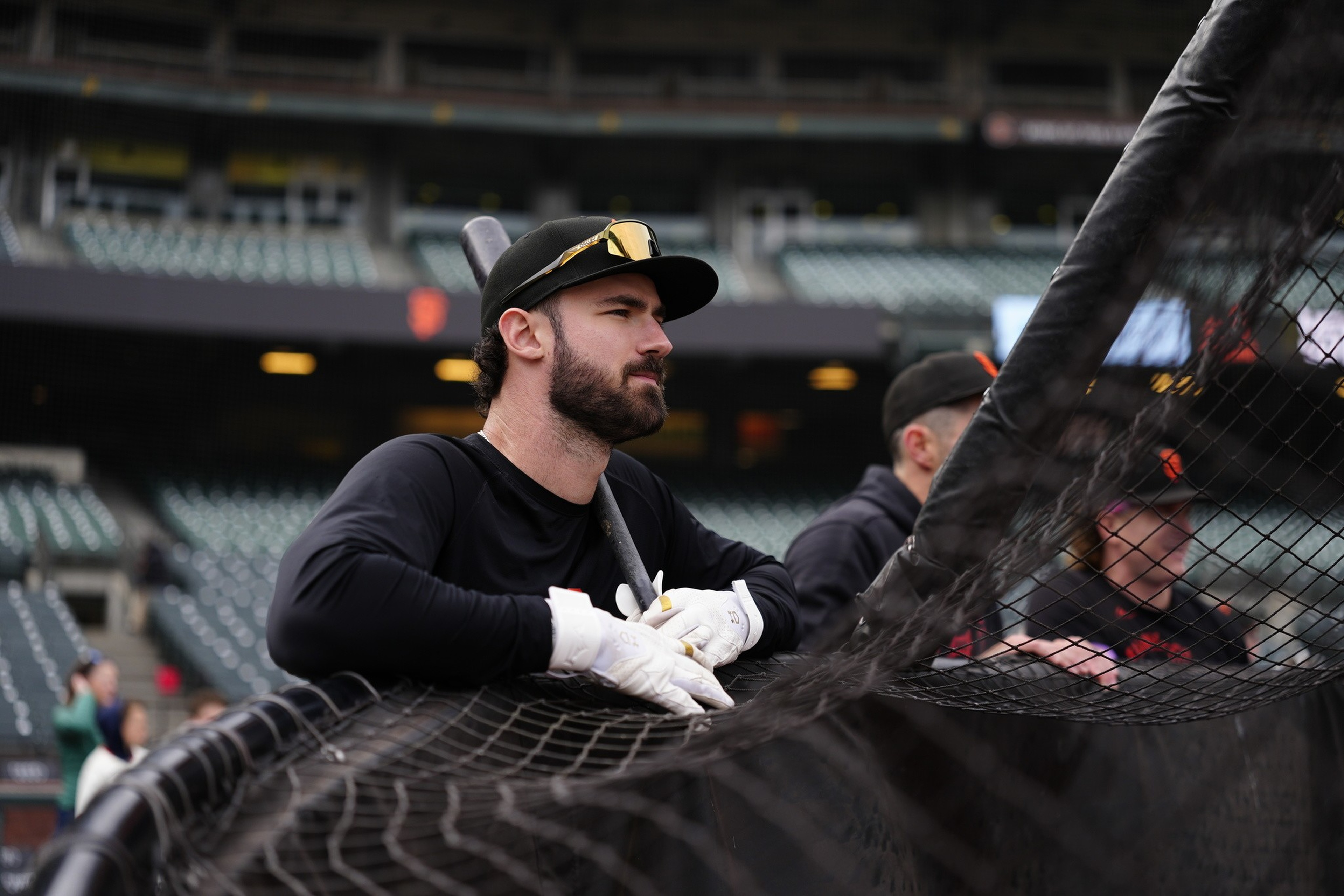 A baseball player wearing black and white gloves leans on the batting cage netting, holding a bat and looking intently ahead in an empty stadium.