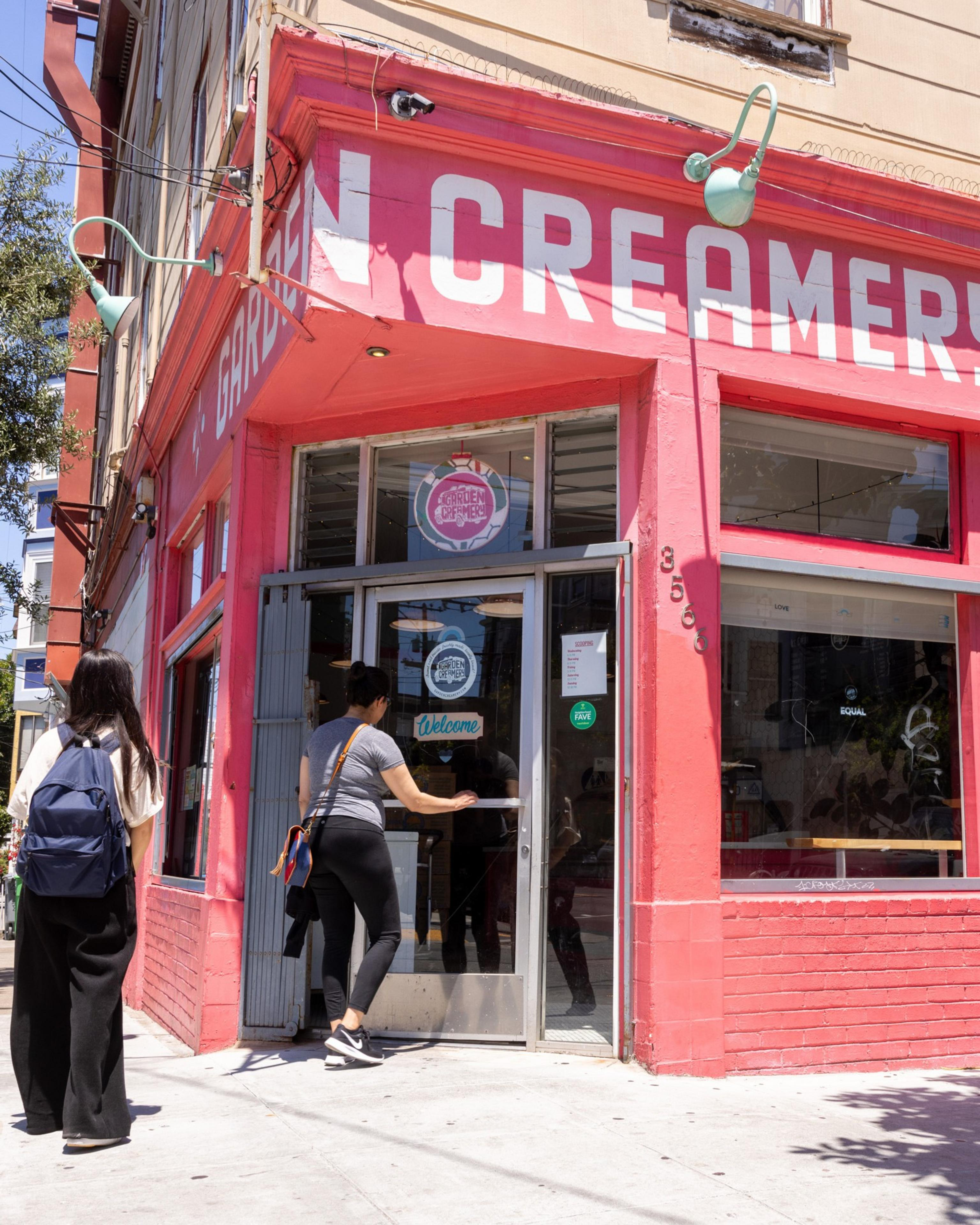 Two people walk into a brightly colored, pink storefront labeled &quot;CREAMERY.&quot; One person opens the door, while the other waits outside with a backpack.