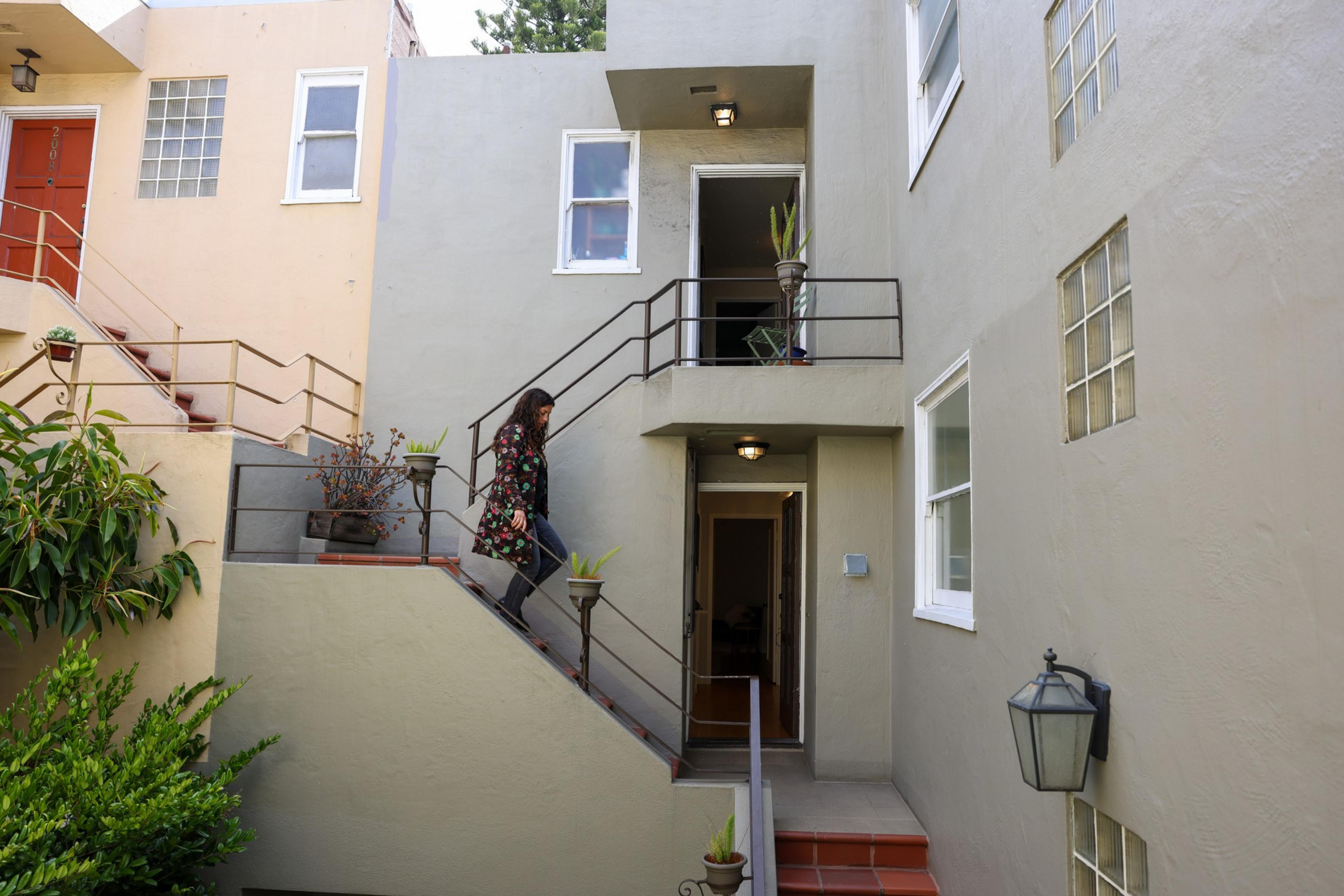 A woman wearing a floral jacket descends an outdoor staircase bordered by potted plants, leading to grey and beige apartment doors.