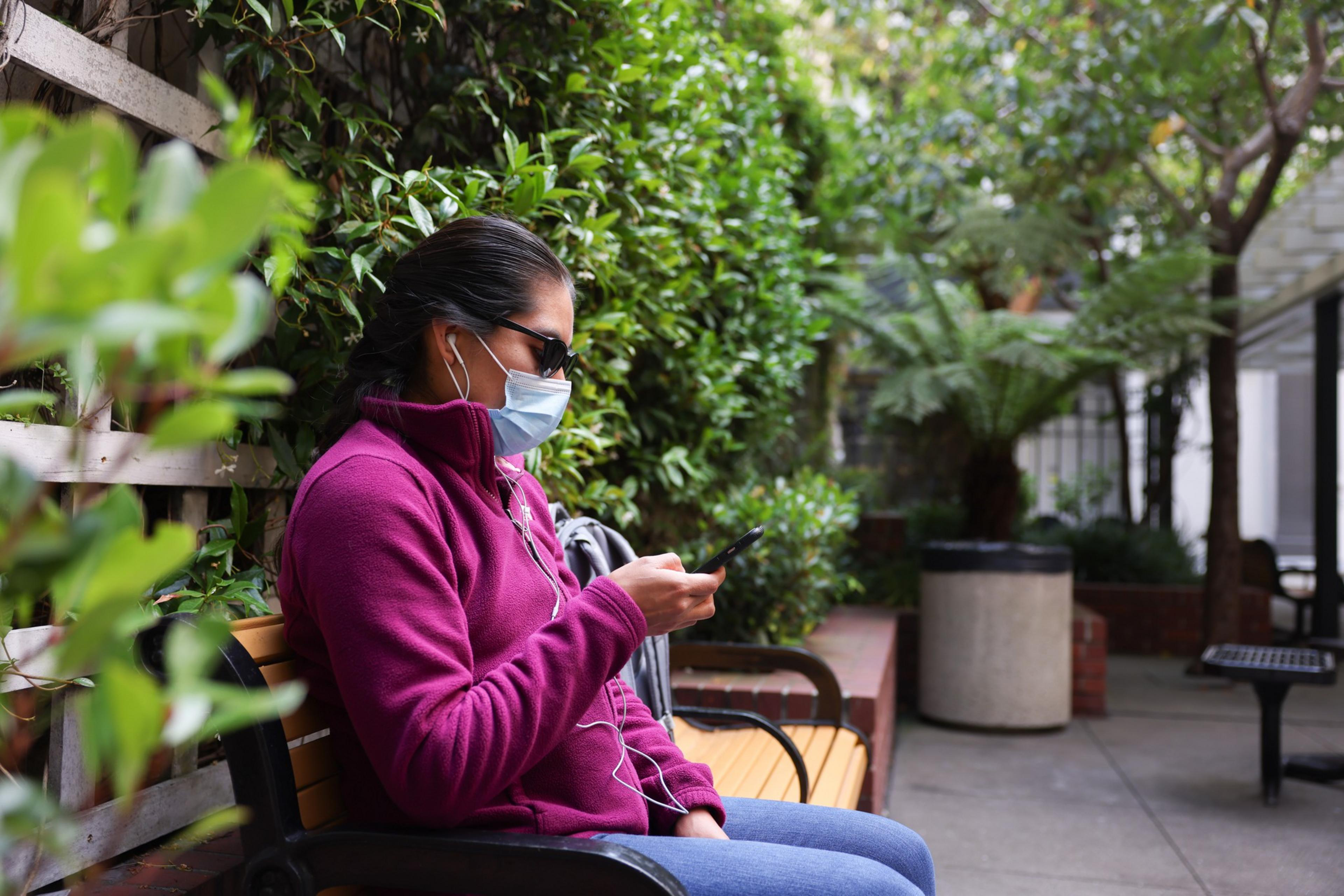 A person wearing a purple jacket, sunglasses, face mask, and earphones sits on a bench outdoors, looking at a smartphone in their hand.