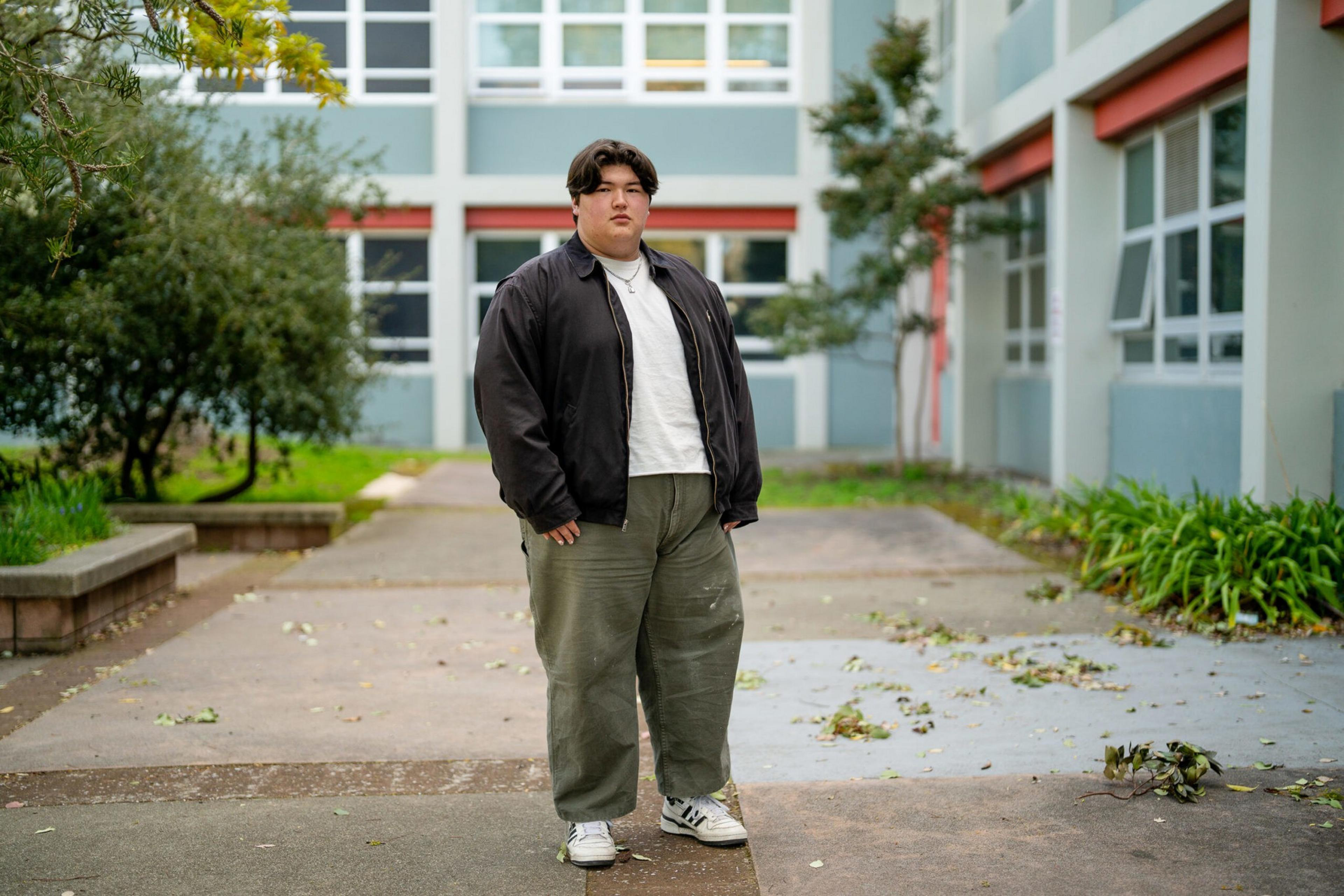 A person wearing a black jacket, white shirt, olive green pants, and white sneakers stands on a sidewalk surrounded by greenery and a blue building.