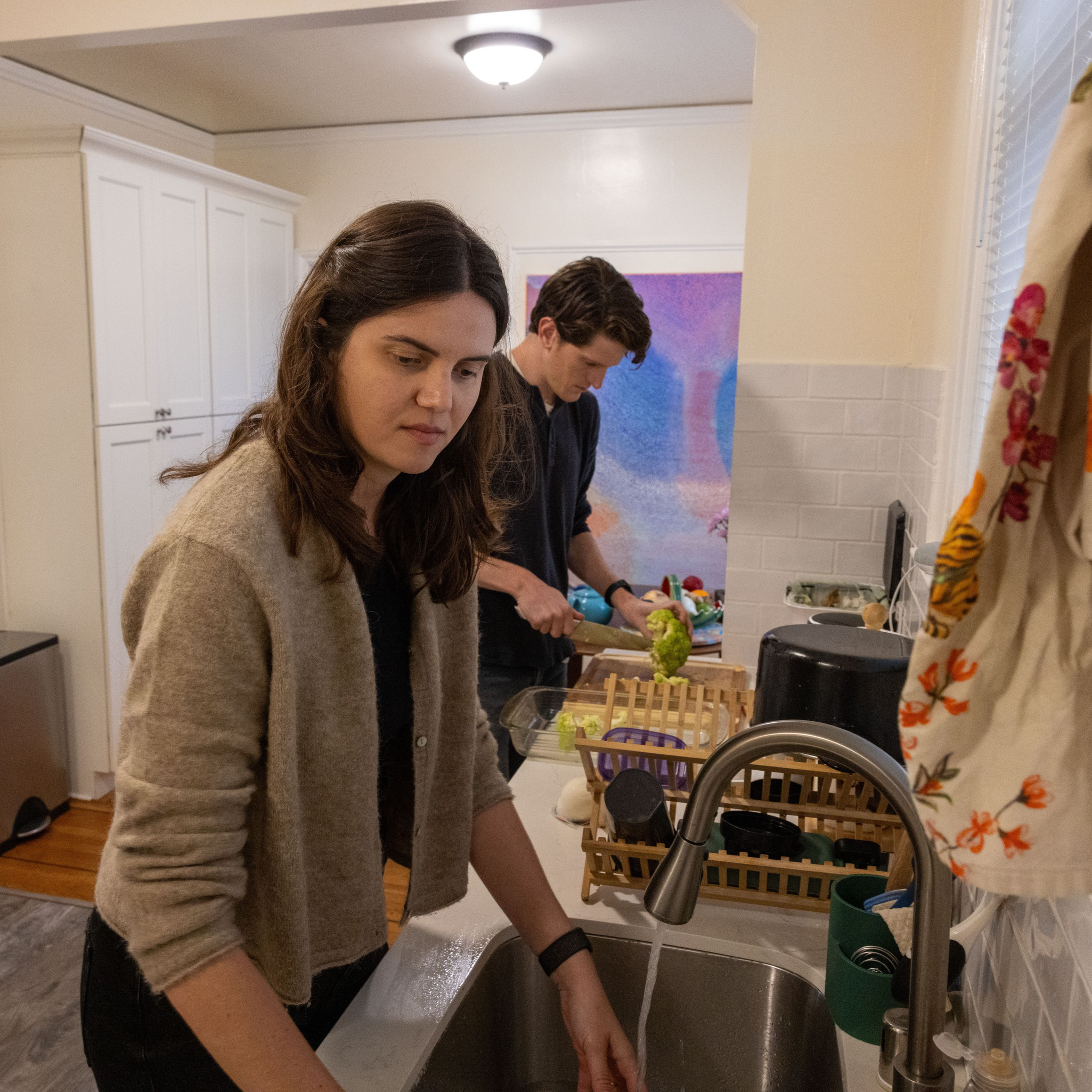 A woman washes dishes at a kitchen sink while a man in the background chops vegetables on a counter. The kitchen has white cabinets and a light beige wall.
