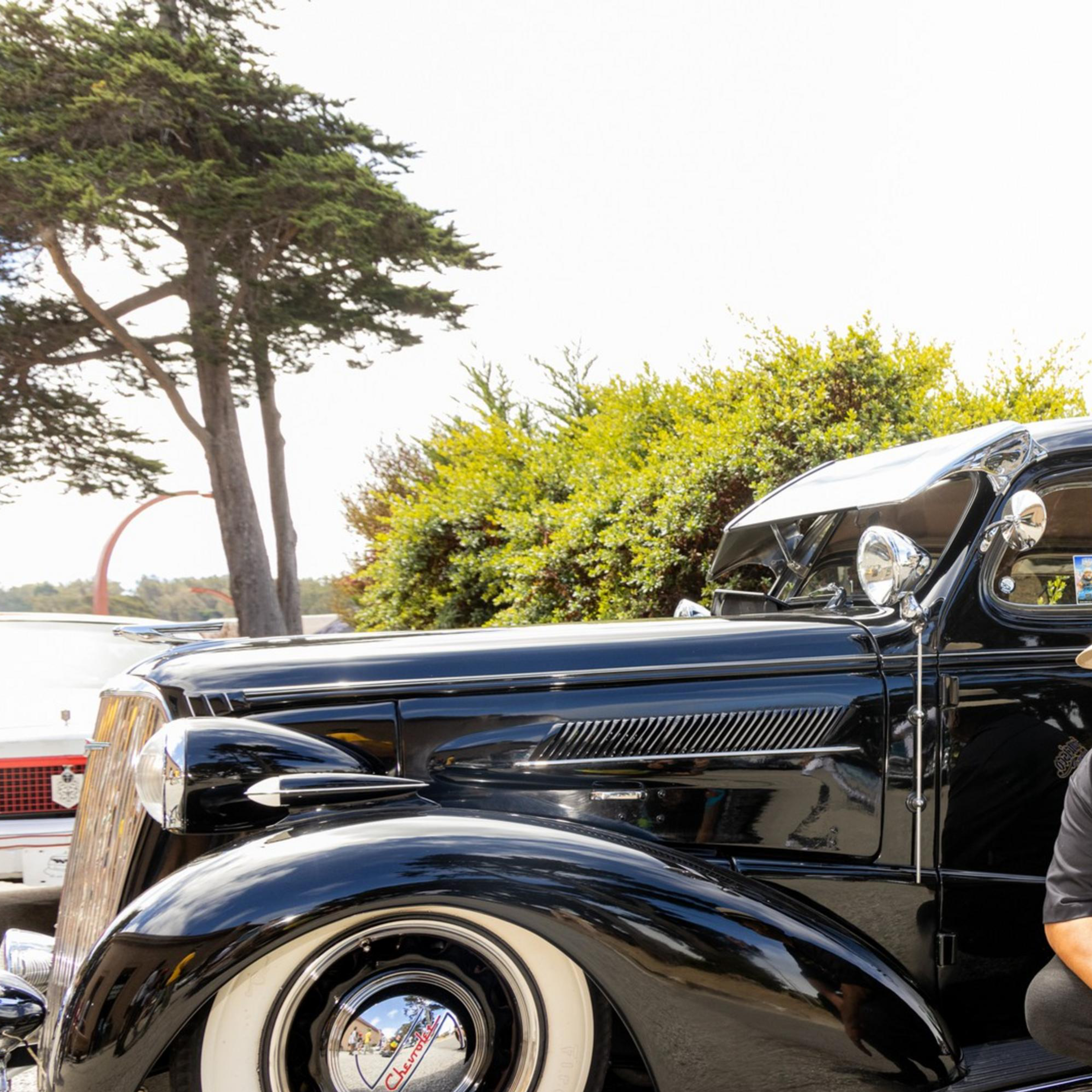 A man kneels beside a polished black vintage car with whitewall tires. He wears a hat, sunglasses, and a black shirt. Trees and other classic cars are in the background.