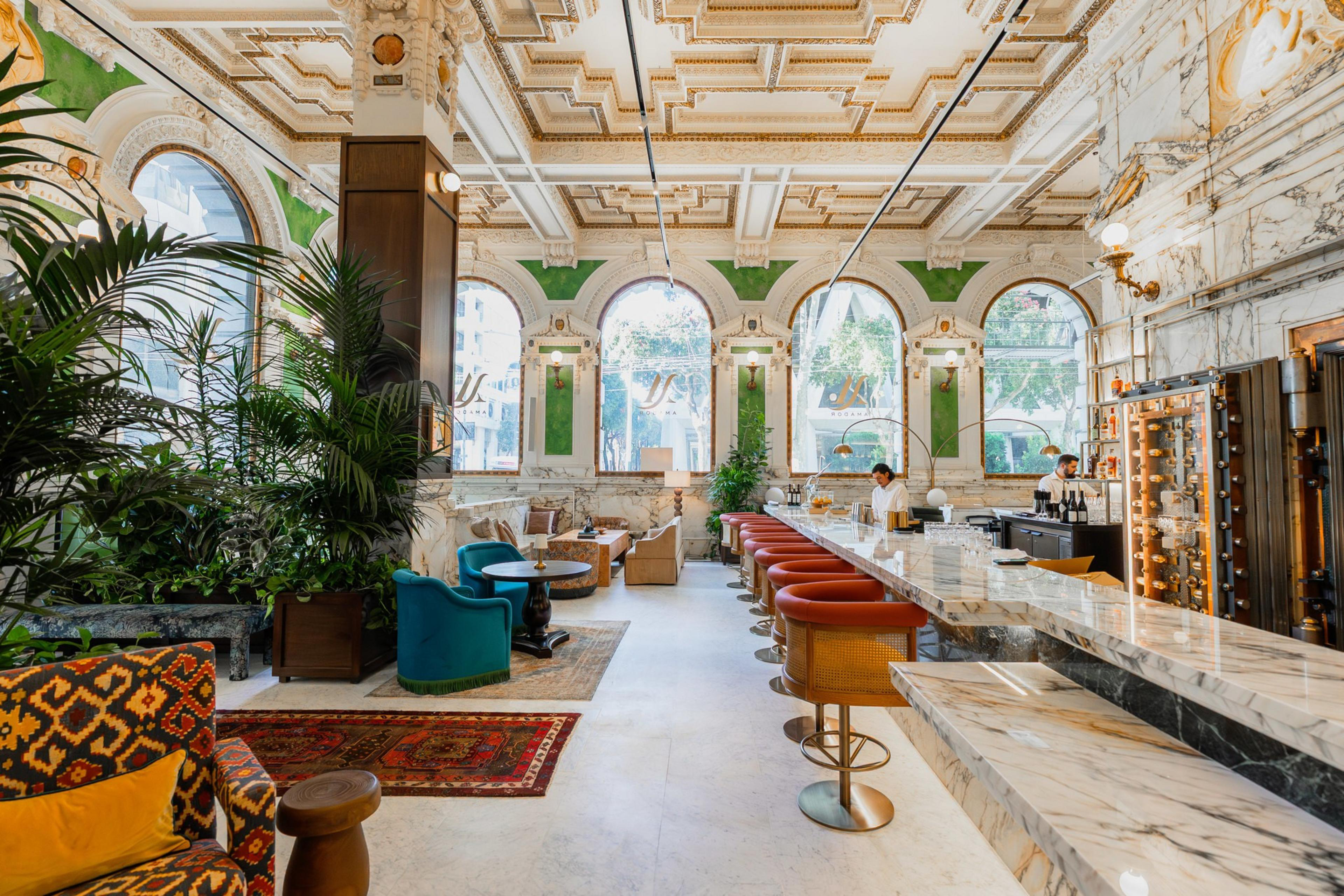 A luxurious bar with a long marble counter, red swivel stools, ornate ceiling, large arched windows, vibrant plants, and comfortable seating areas.
