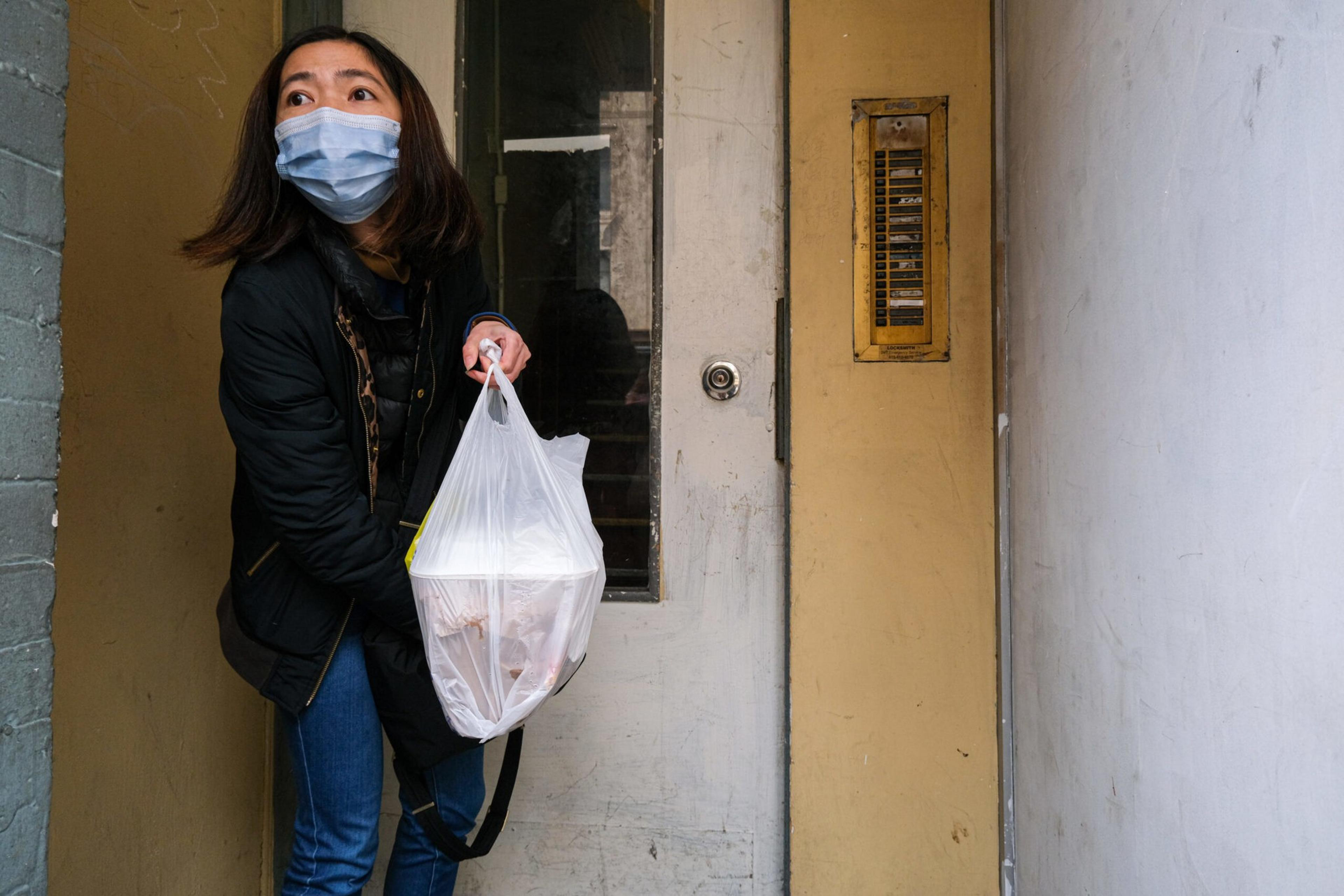 A woman wearing a face mask and black jacket holds a plastic bag with food while standing in front of a door in a narrow hallway.