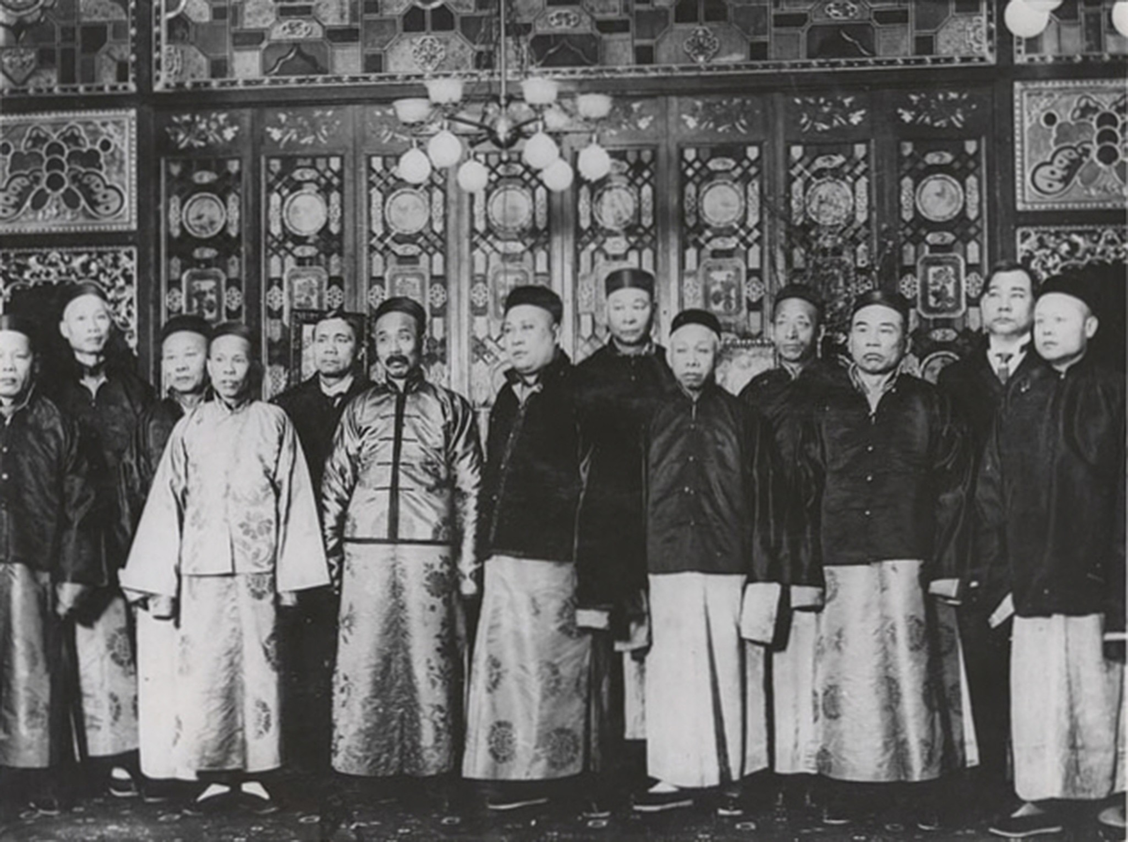 A group of fourteen men in traditional Chinese robes stand in front of an ornate, decorated wall and a chandelier.