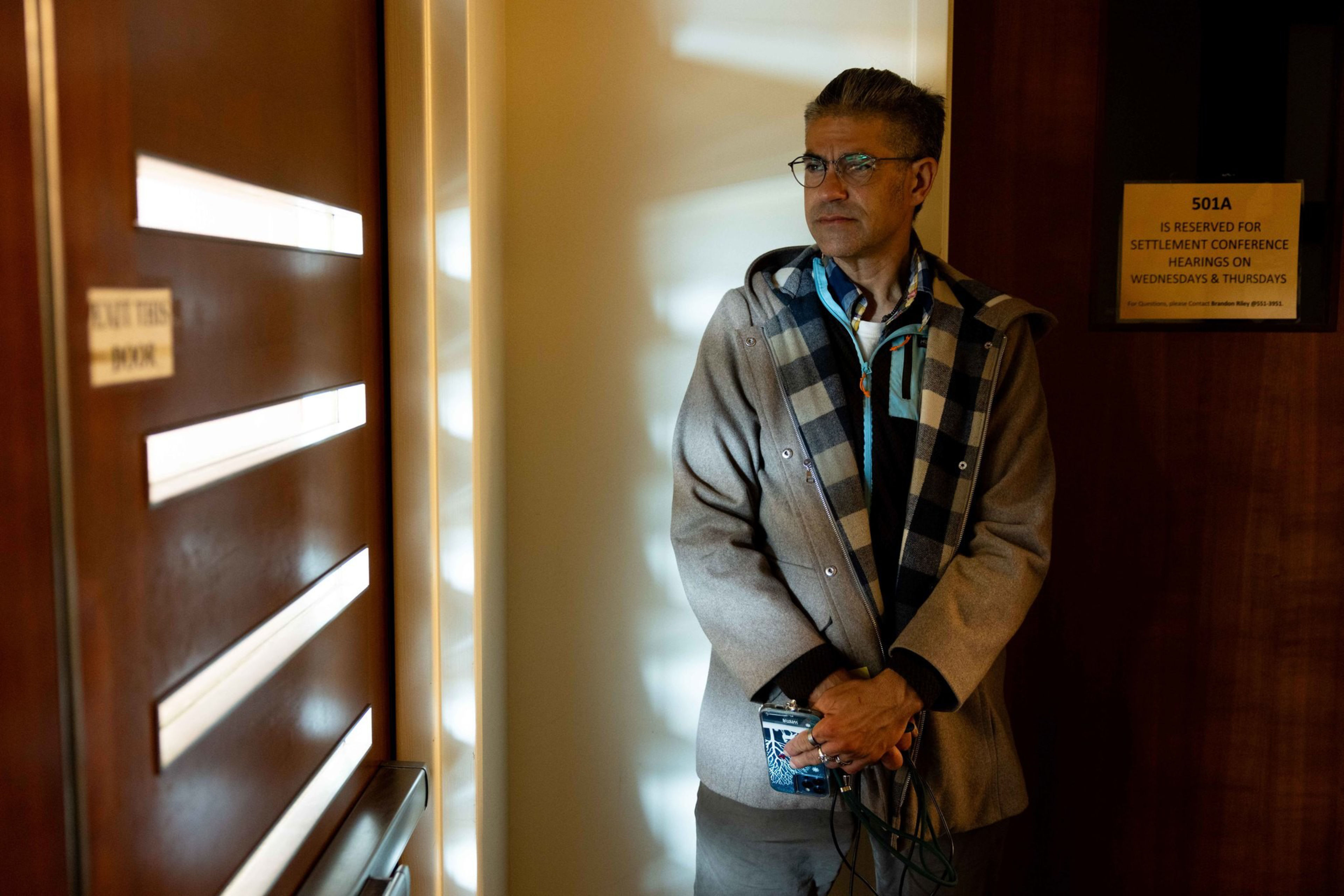 A man wearing glasses, a gray coat, and a plaid scarf stands against a wall holding a blue cup and black cables near a room labeled 501A.
