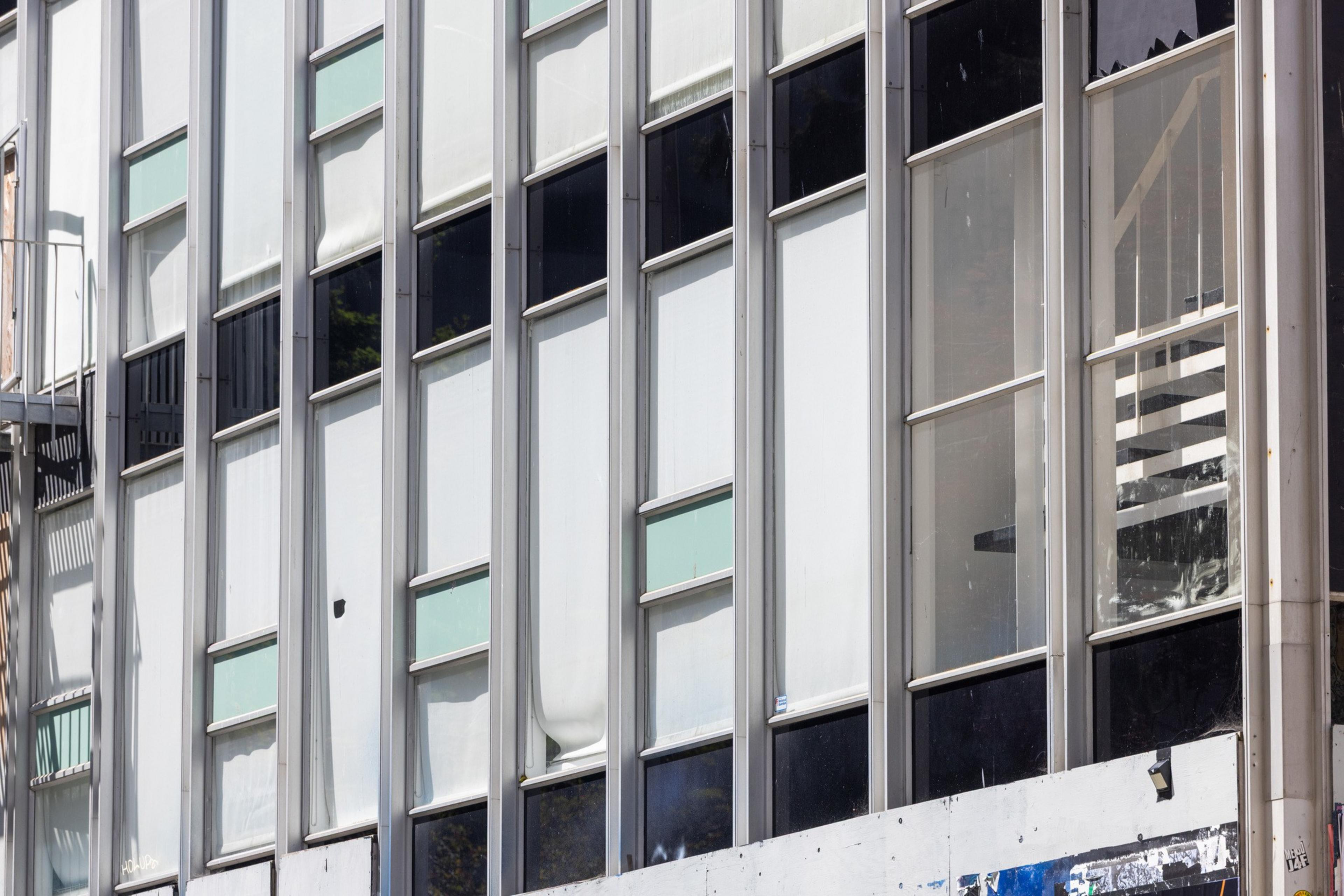 The image shows a building facade with large vertical windows framed in metal. Some windows have blinds partially drawn, and a fire escape stairs are visible on the left.