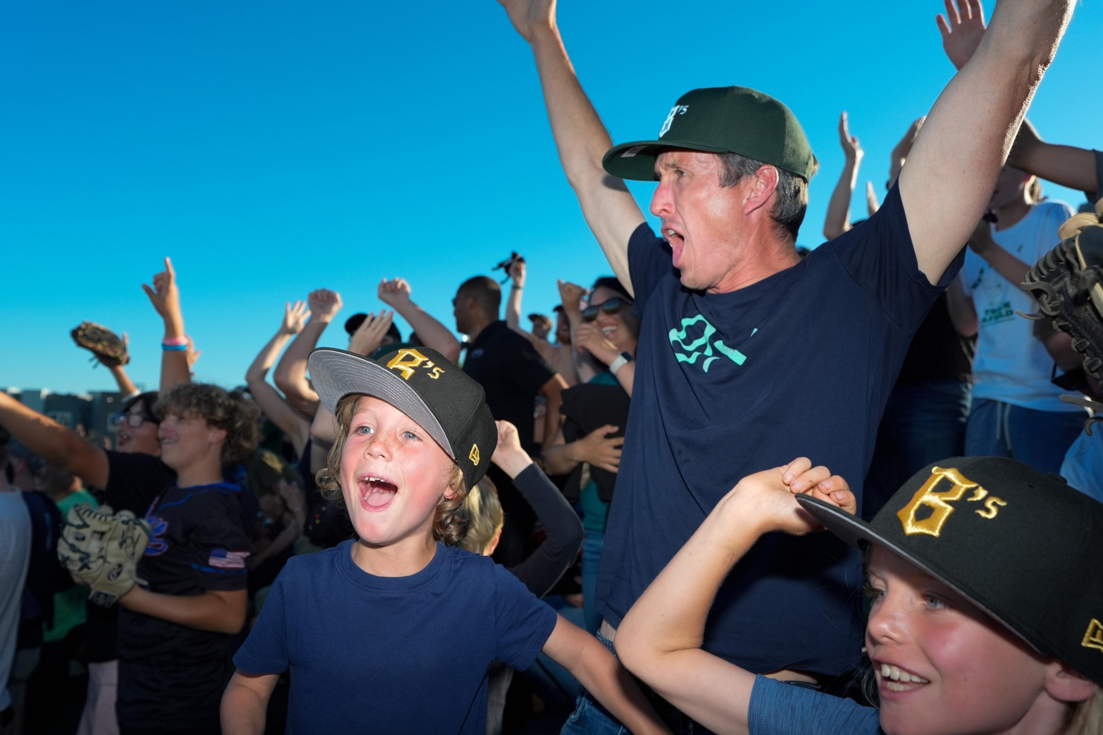 The image shows excited fans, including children and adults, cheering with their hands in the air. They are wearing matching team hats and appear to be at a sporting event.
