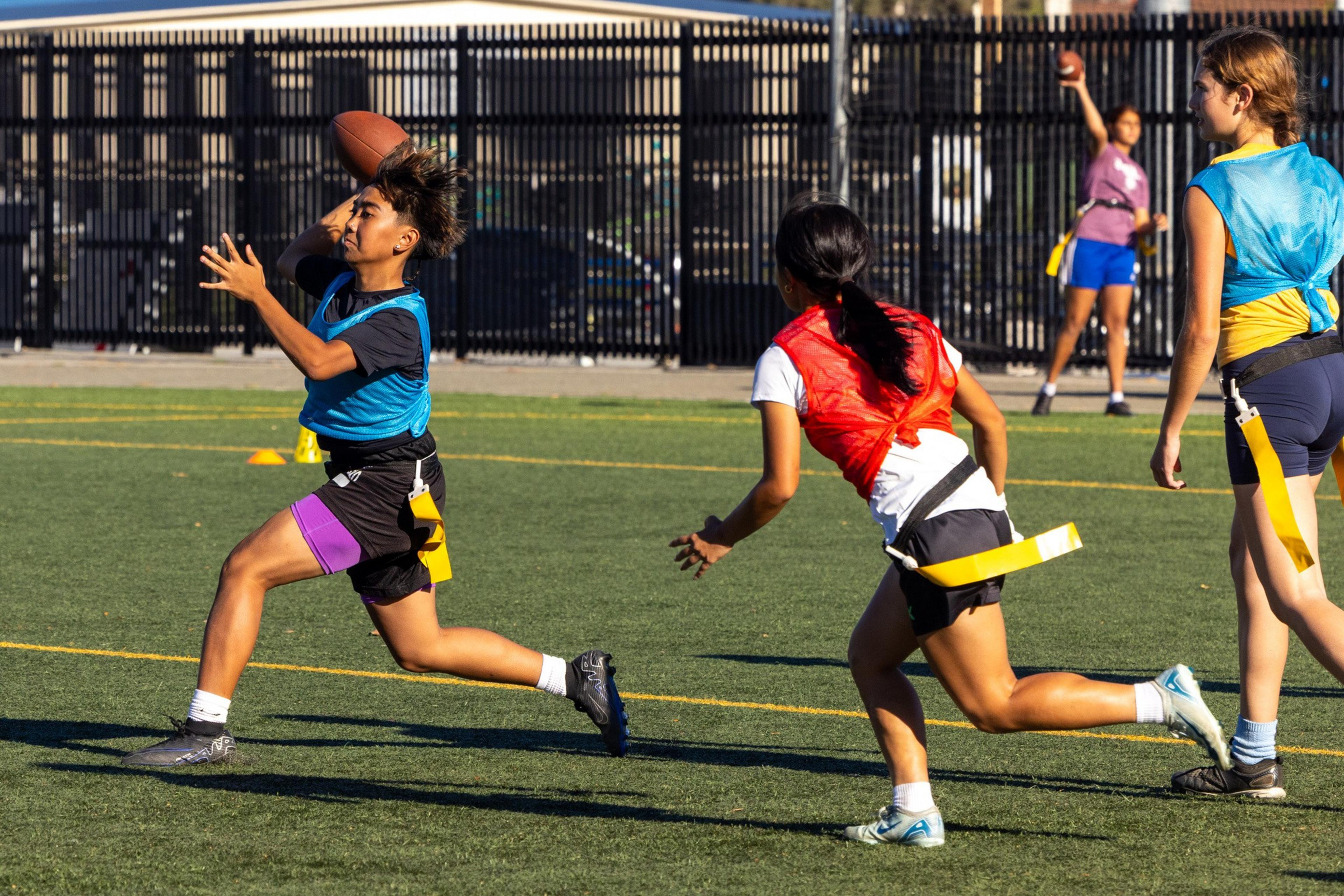 A girl in a blue jersey prepares to catch a football while a girl in a red jersey runs toward her on a grassy field, with two other players in the background.