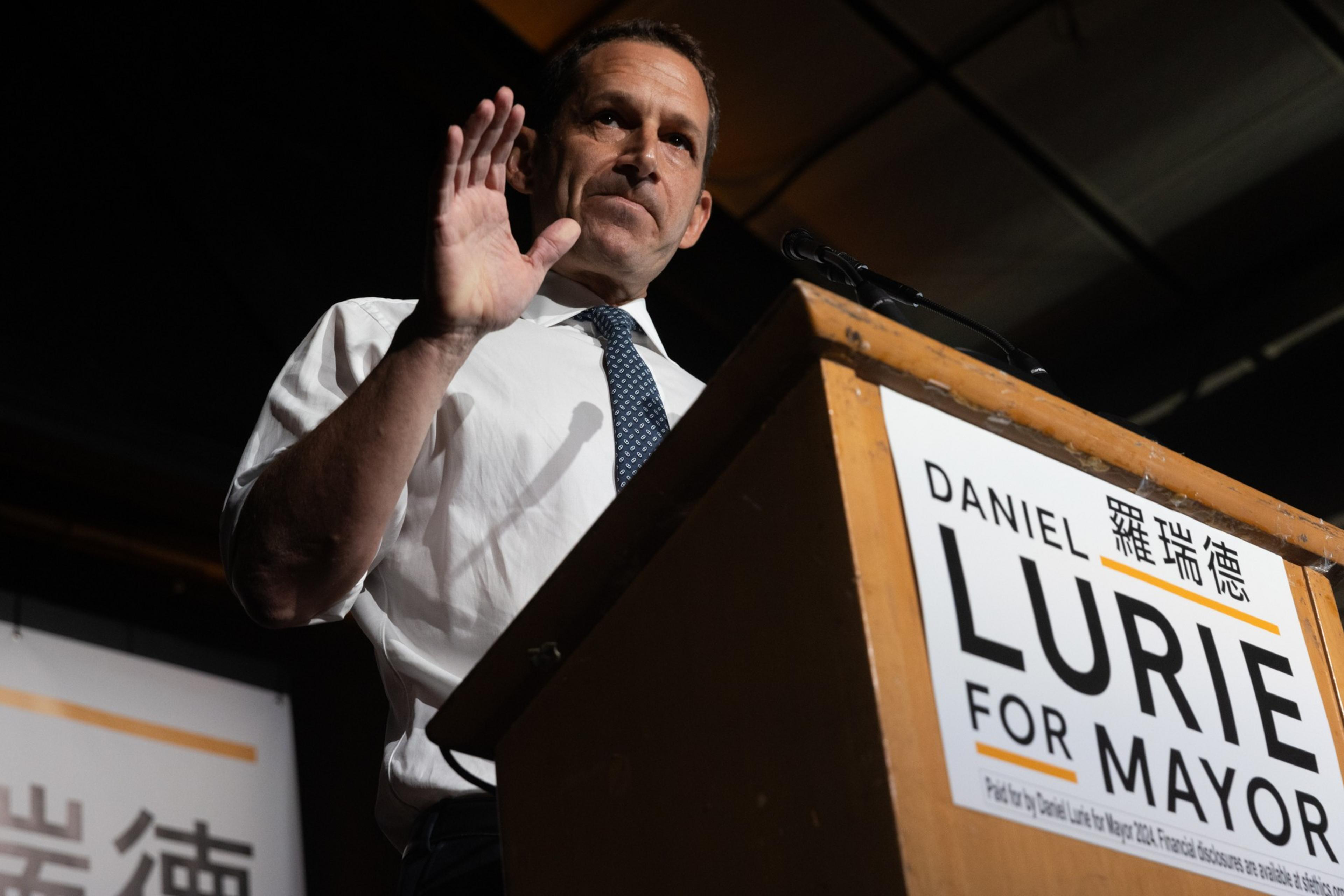 A man in a white shirt and tie gestures while speaking at a podium labeled &quot;FOR MAYOR.&quot;