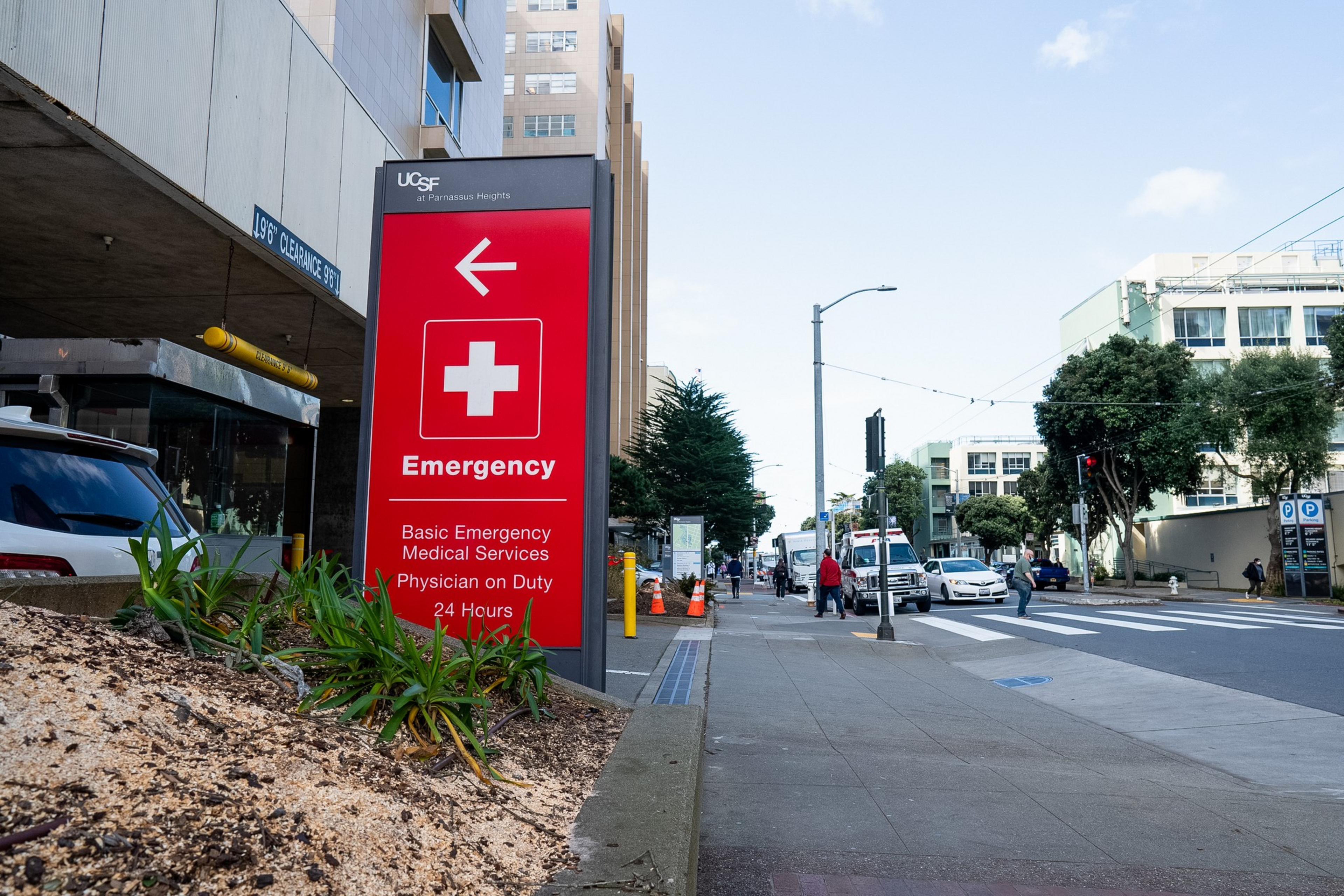 A red emergency sign with a white cross and arrow points left. The street has an ambulance, cars, pedestrians, and trees. Buildings line the background.