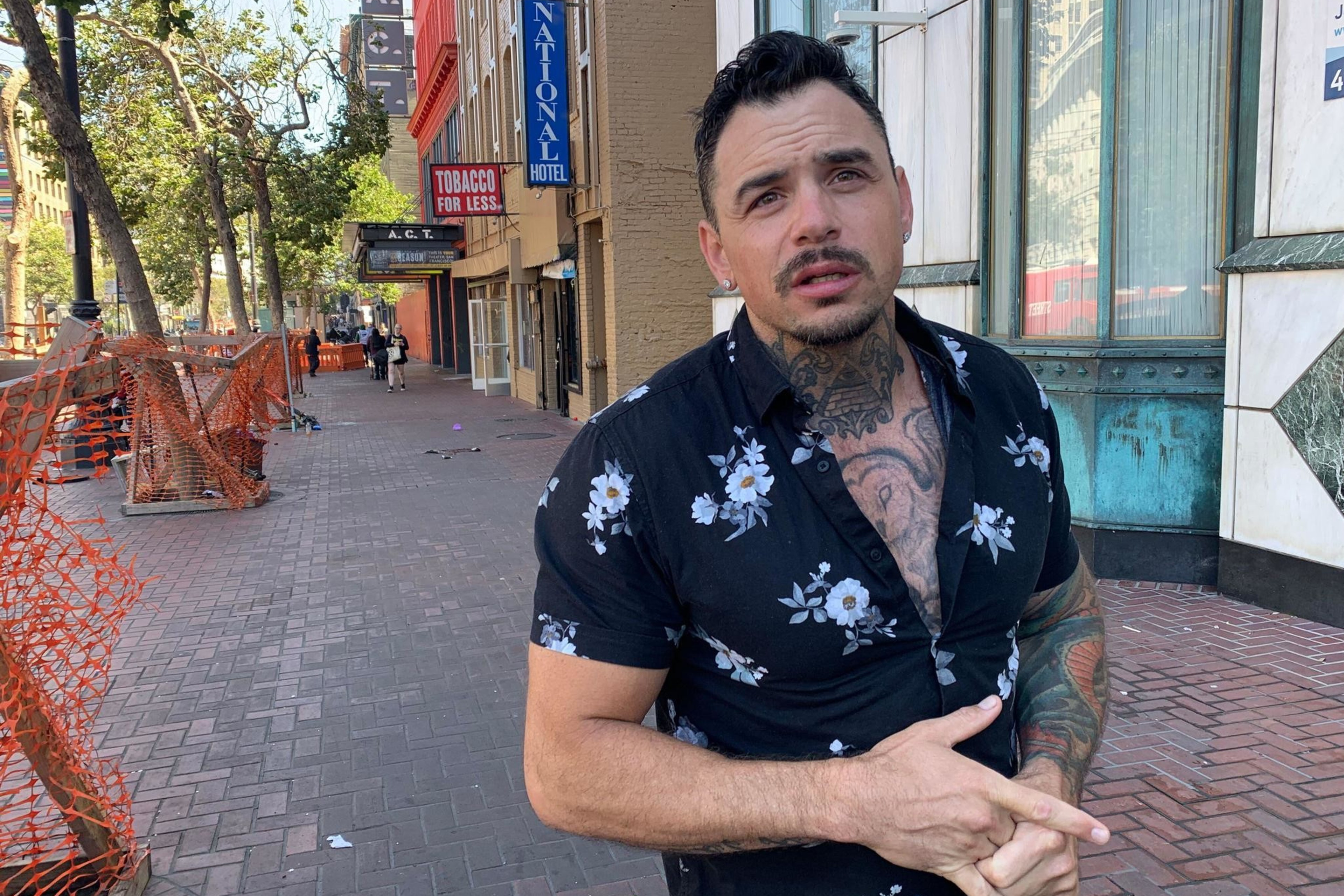 A man with tattoos, wearing a floral shirt, stands on a brick sidewalk. Behind him are trees, orange fencing, and storefronts, including a ""Tobacco for Less"" sign.