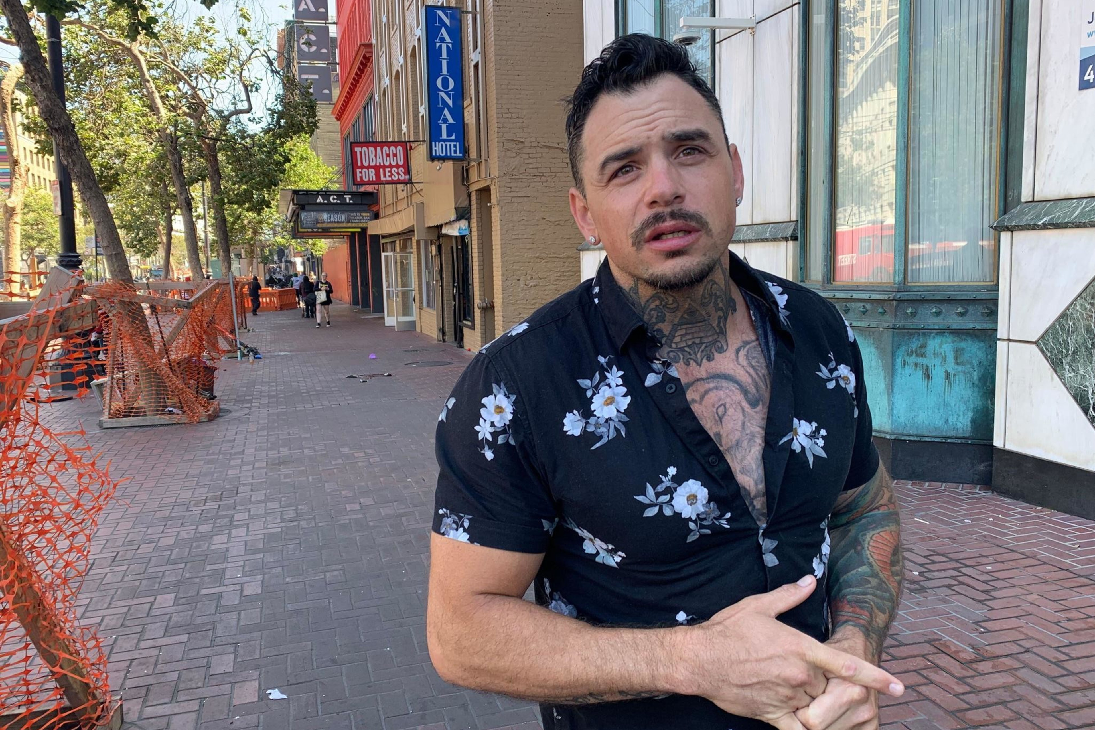 A man with tattoos, wearing a floral shirt, stands on a brick sidewalk. Behind him are trees, orange fencing, and storefronts, including a ""Tobacco for Less"" sign.