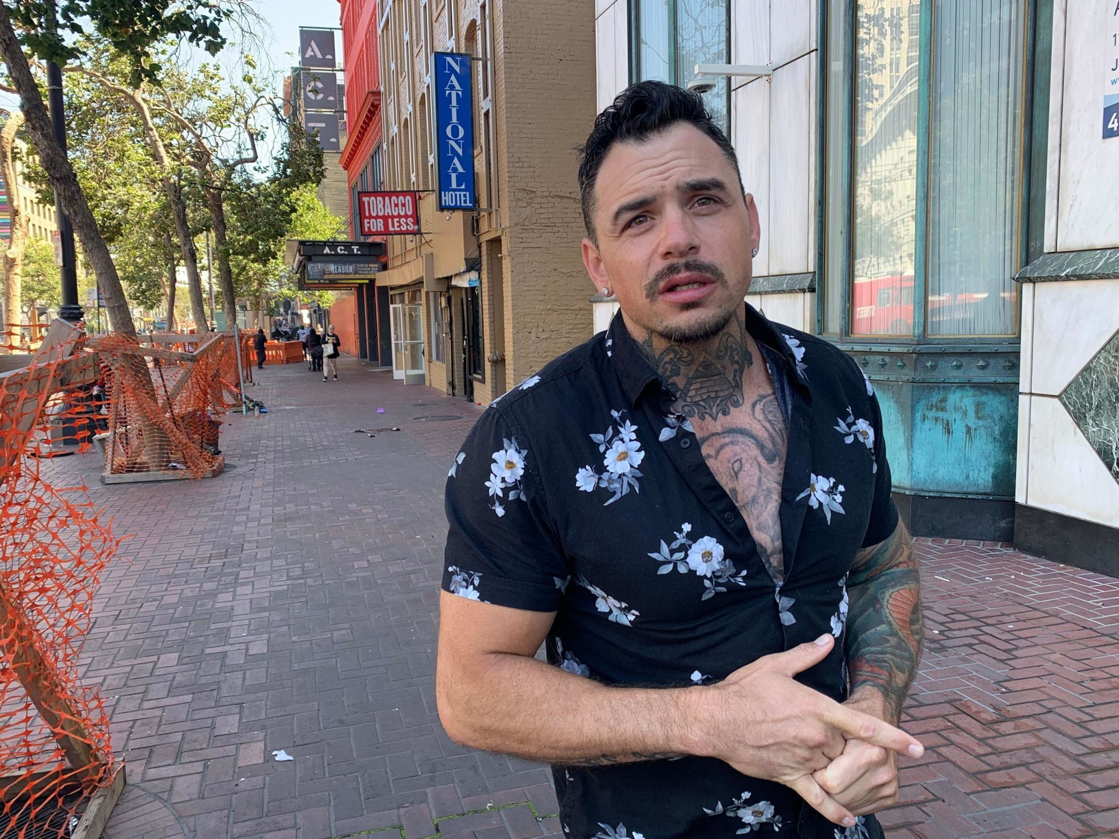 A man with tattoos, wearing a floral shirt, stands on a brick sidewalk. Behind him are trees, orange fencing, and storefronts, including a ""Tobacco for Less"" sign.