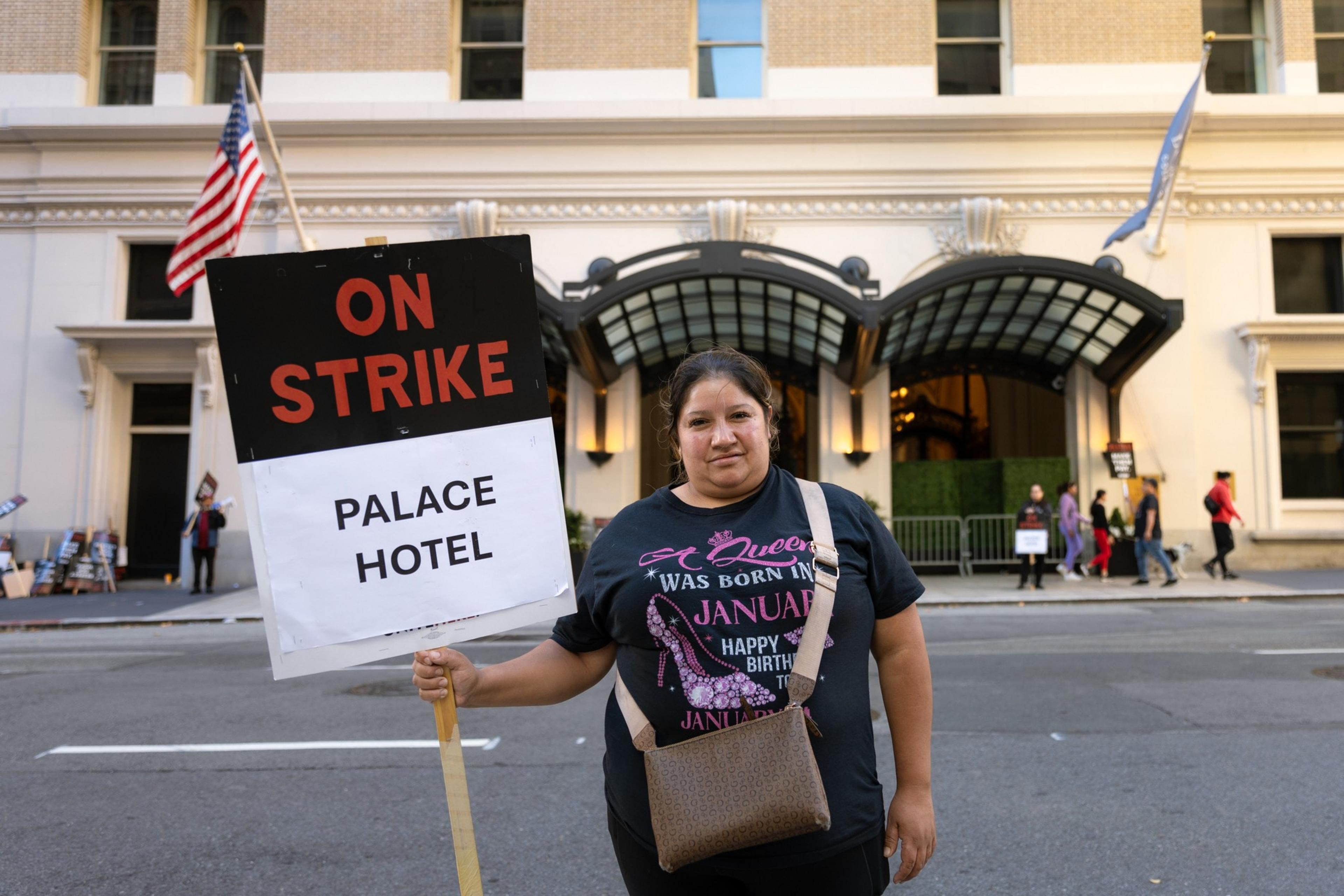 A woman in a black shirt stands in front of a hotel holding a sign reading &quot;ON STRIKE: PALACE HOTEL.&quot; An American flag is seen in the background.