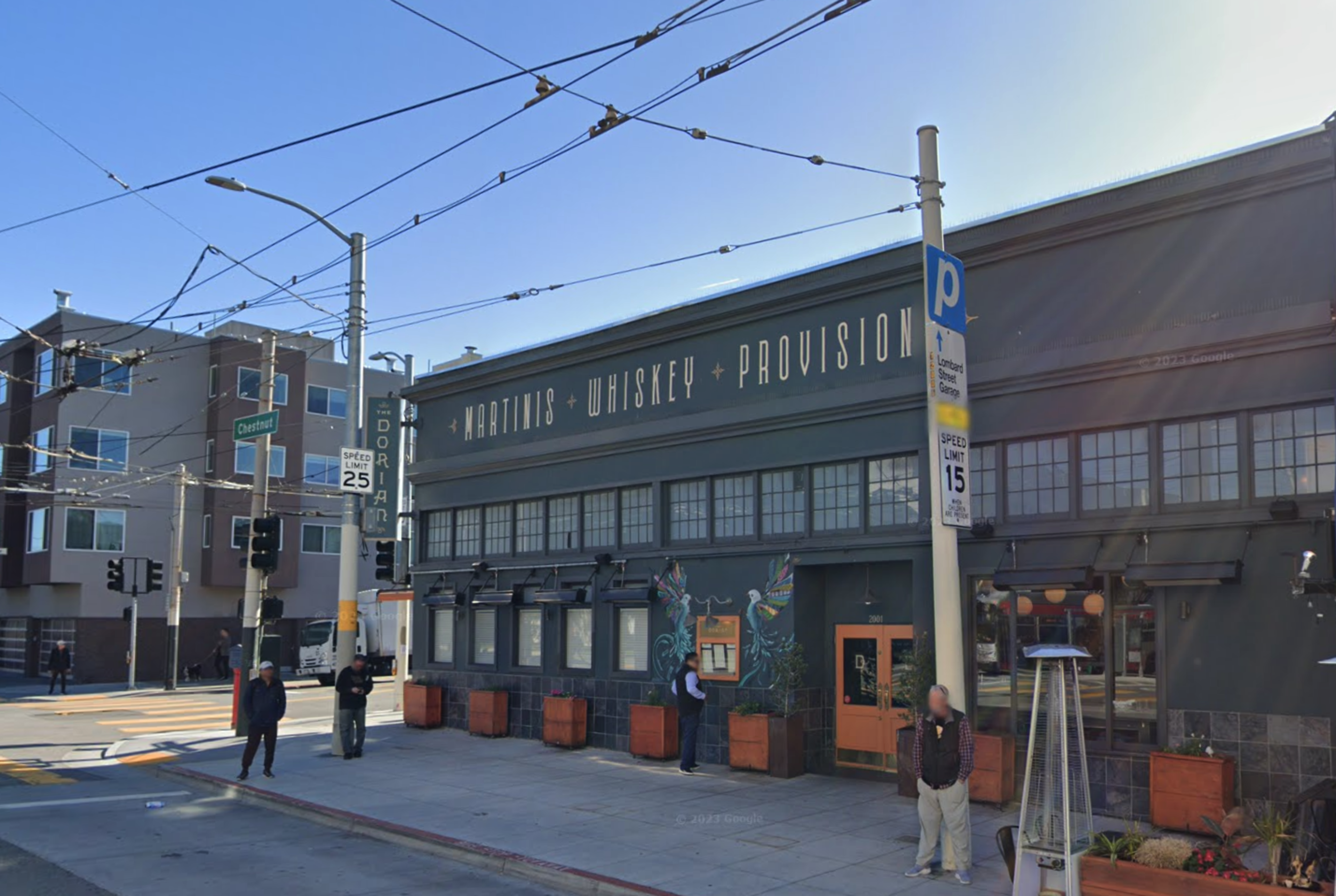 A dark gray building with "Martinis Whiskey Provision" written on it, located on a street corner with several people standing on the sidewalk nearby.