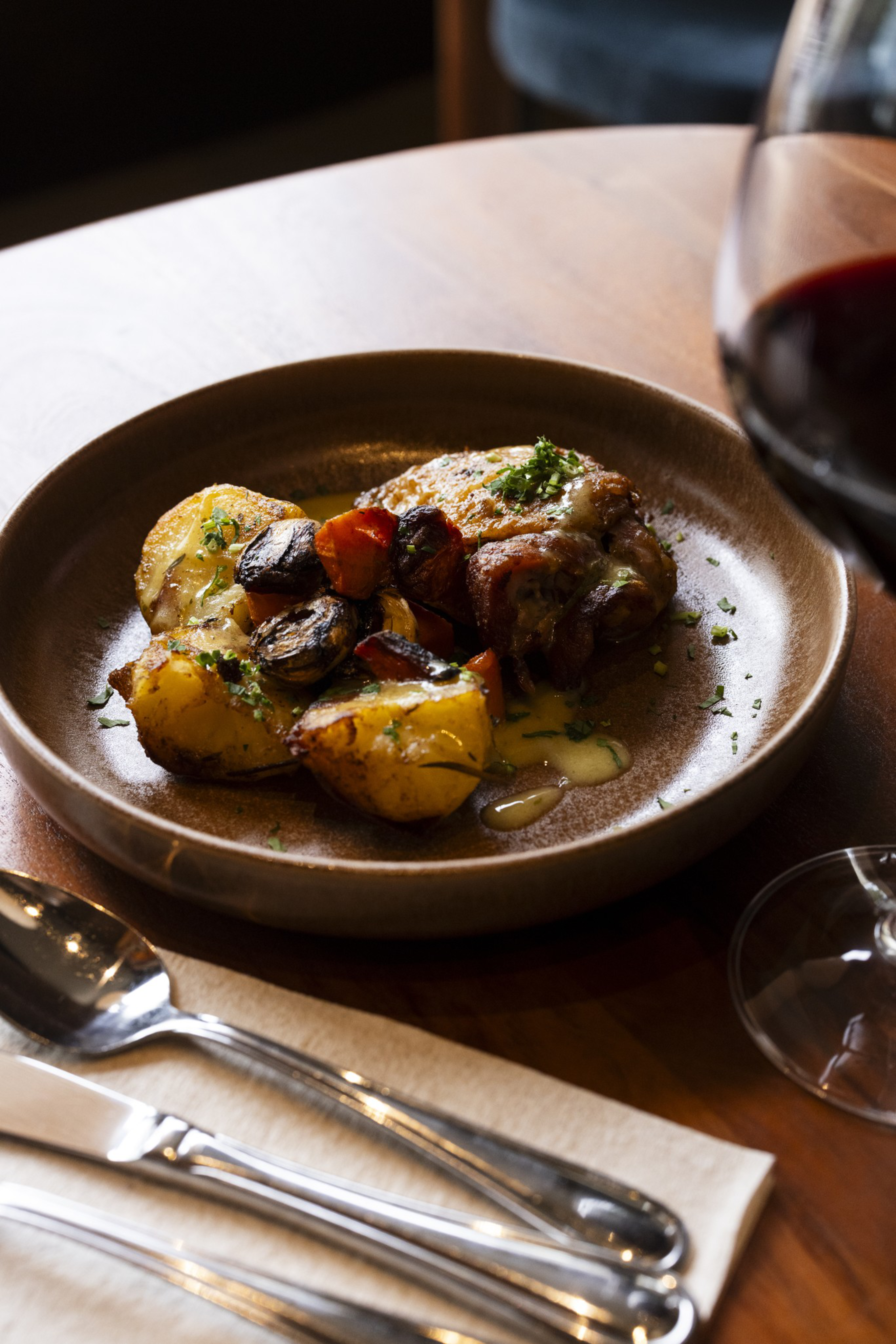 Roasted chicken with golden potatoes and mushrooms garnished with herbs, served on a brown plate next to a glass of red wine and silver cutlery.