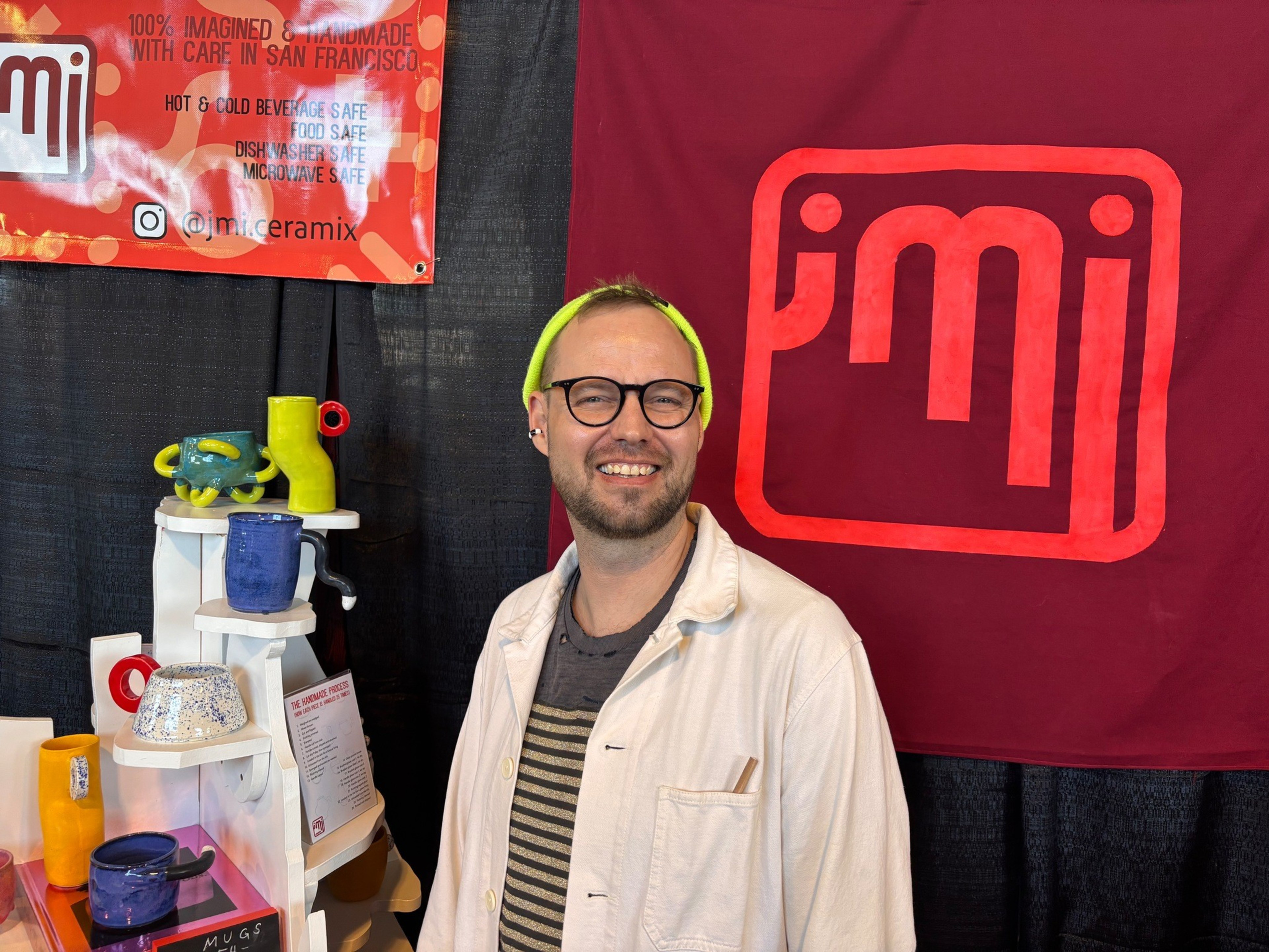 A smiling person with glasses and a neon yellow headband stands beside a display of colorful ceramic mugs and a red banner with the “jmi” logo.
