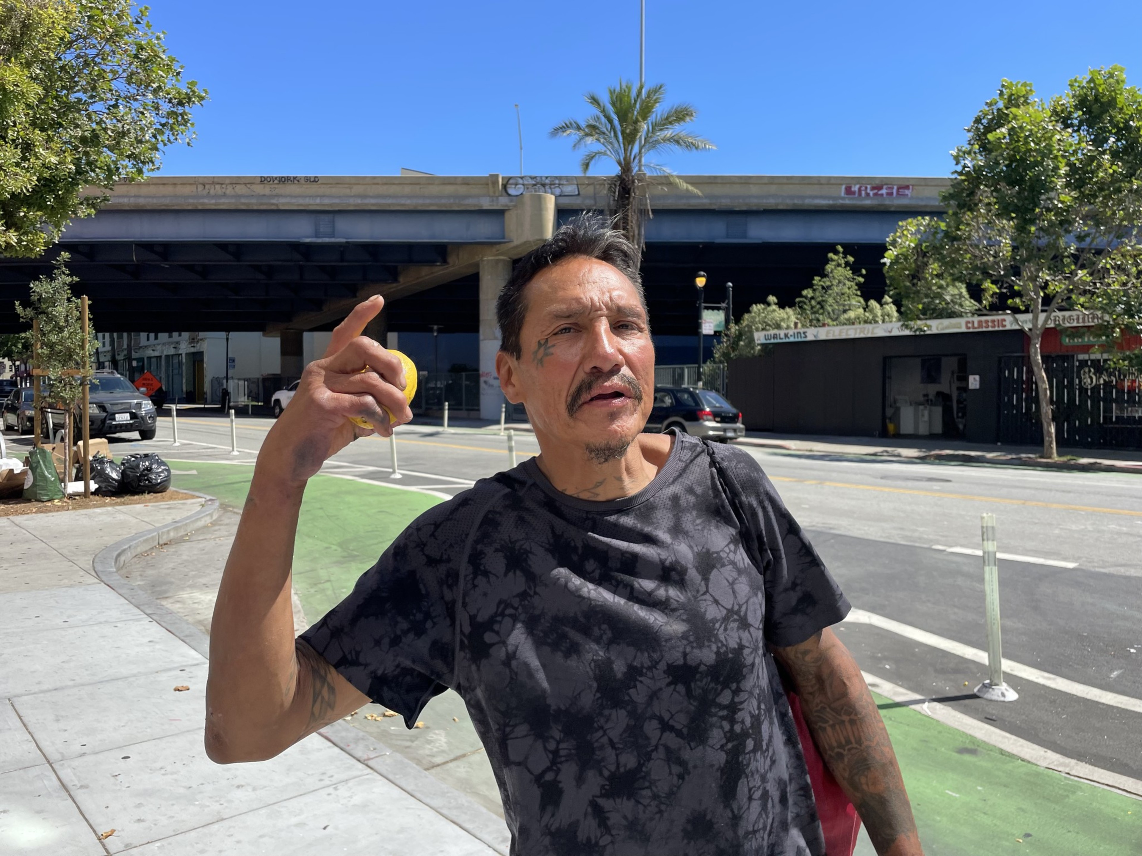 A man with tattoos, wearing a black patterned shirt, holds up a yellow object while standing outside with a bridge, palm tree, and shops in the background on a sunny day.