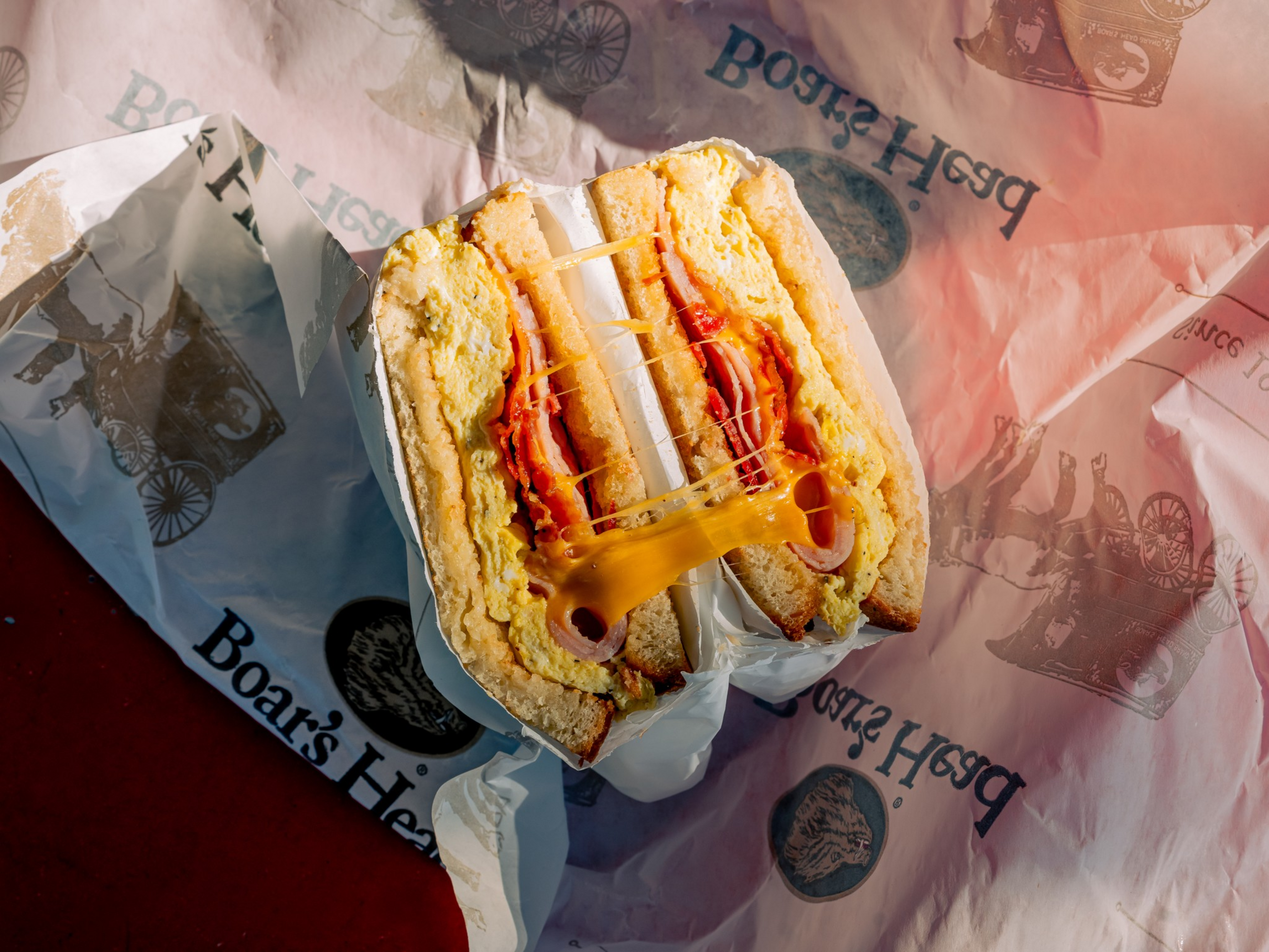 A sandwich cut in half with melted cheese, scrambled eggs, and slices of ham between pieces of bread, resting on branded paper wrapping.