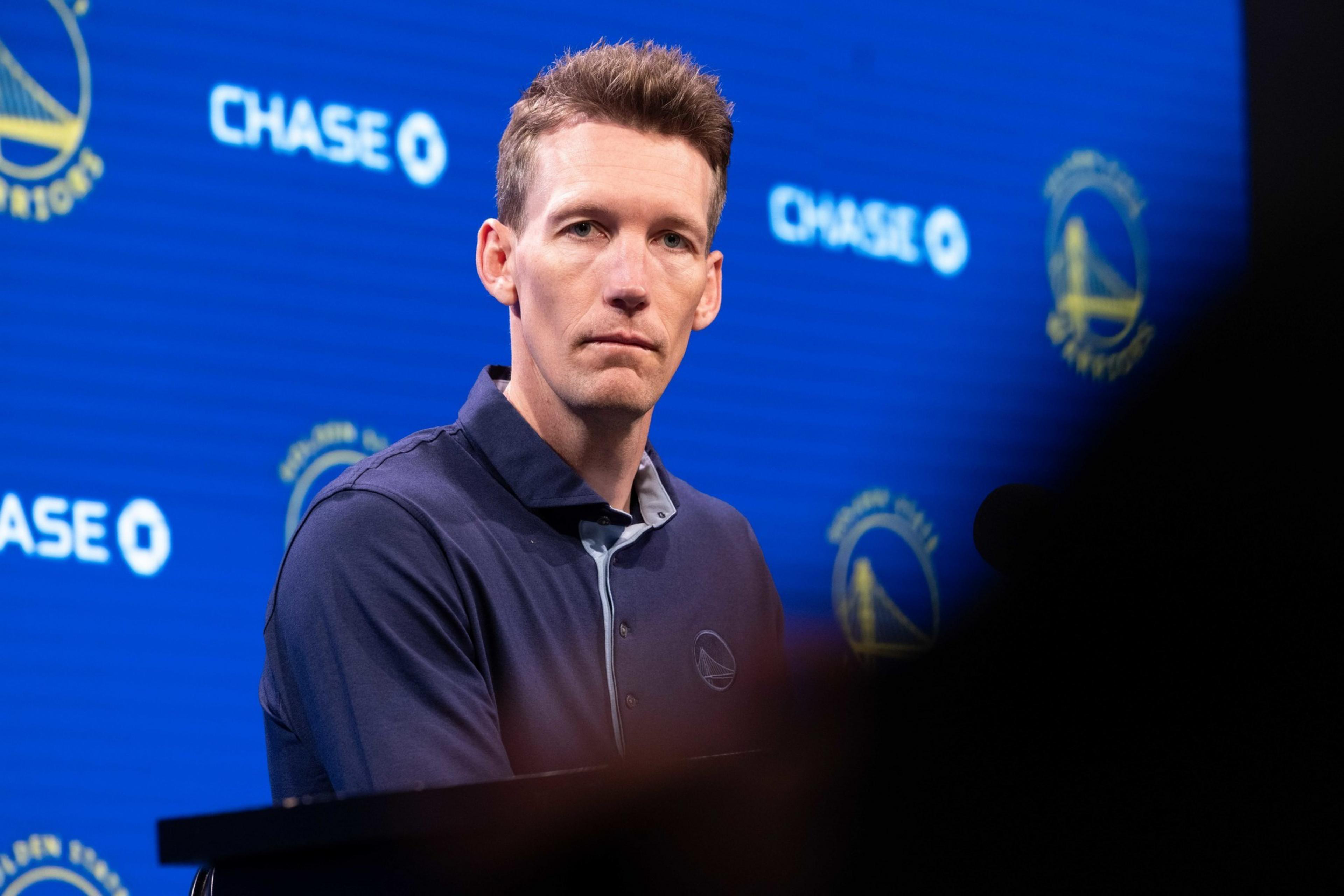 A man in a dark collared shirt sits in front of a blue background featuring logos and &quot;CHASE&quot; text, looking serious or focused.
