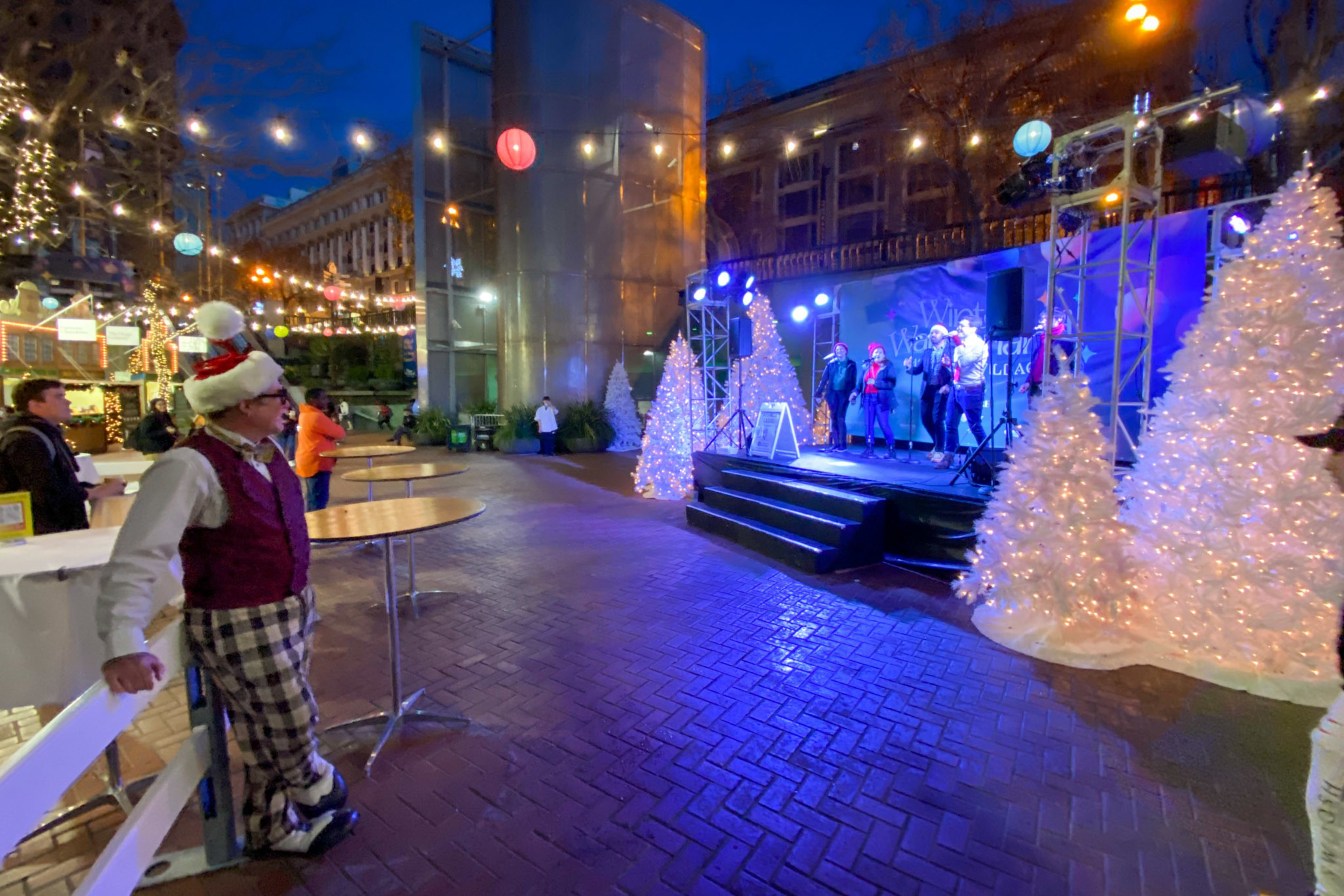 A man in Christmas attire watches singers performing on a stage decorated with white lit Christmas trees and festive lights in an outdoor plaza at night.
