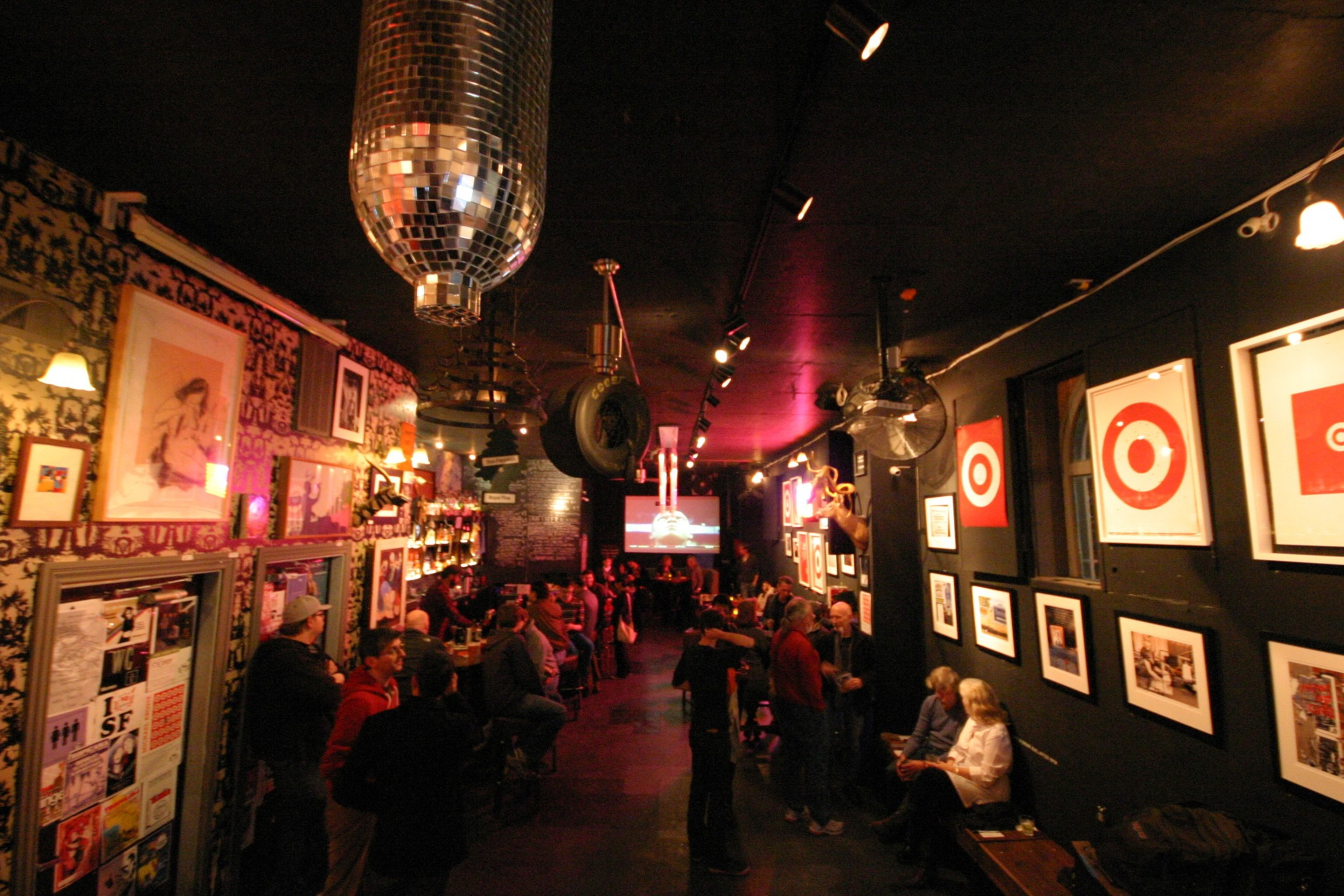 A dimly lit bar with people socializing, walls covered in framed art, and a large disco ball hanging from the ceiling.