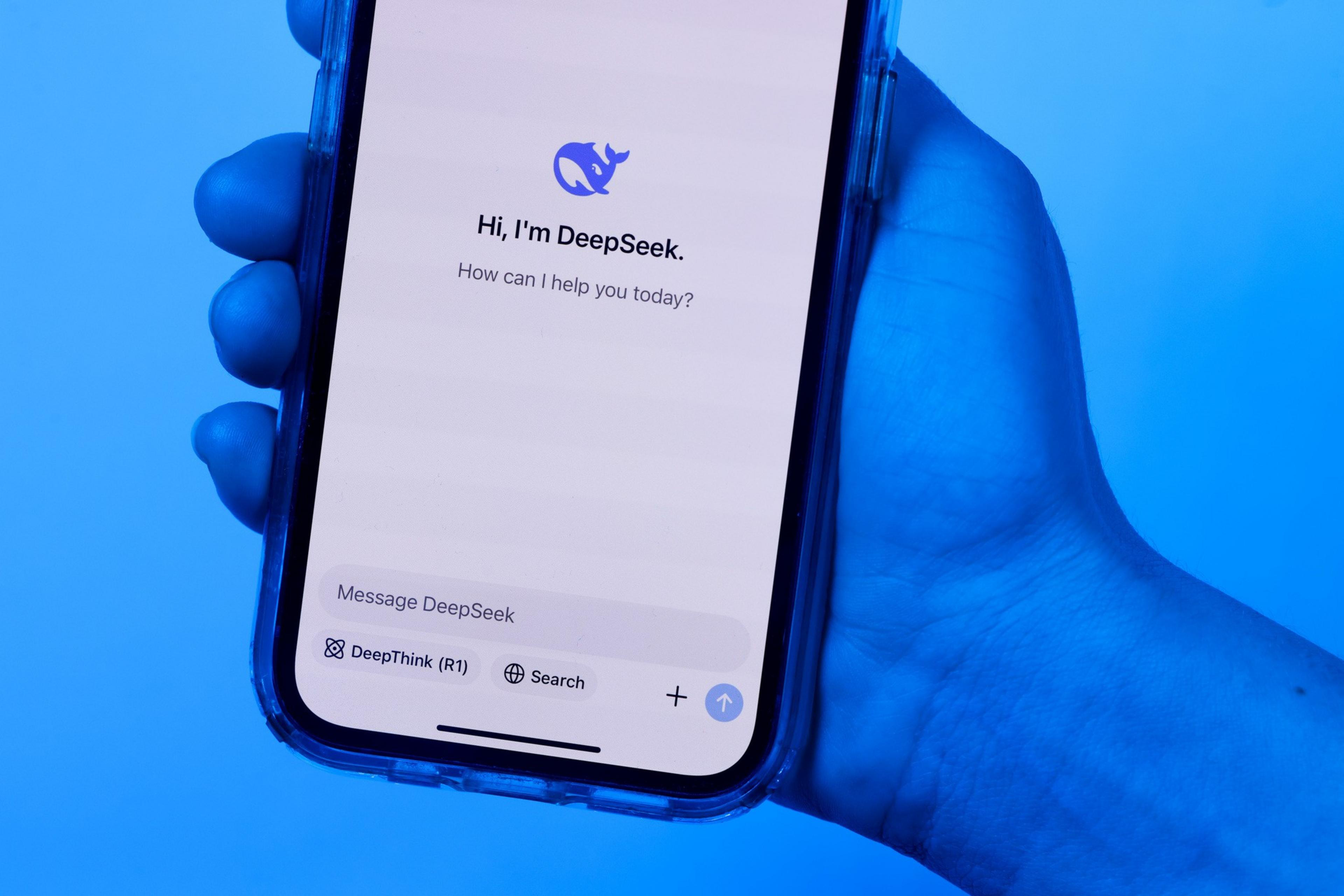 A hand holds a phone displaying a screen with "Hi, I'm DeepSeek. How can I help you today?" and options for messaging, DeepThink, and Search. The image has a blue tint.